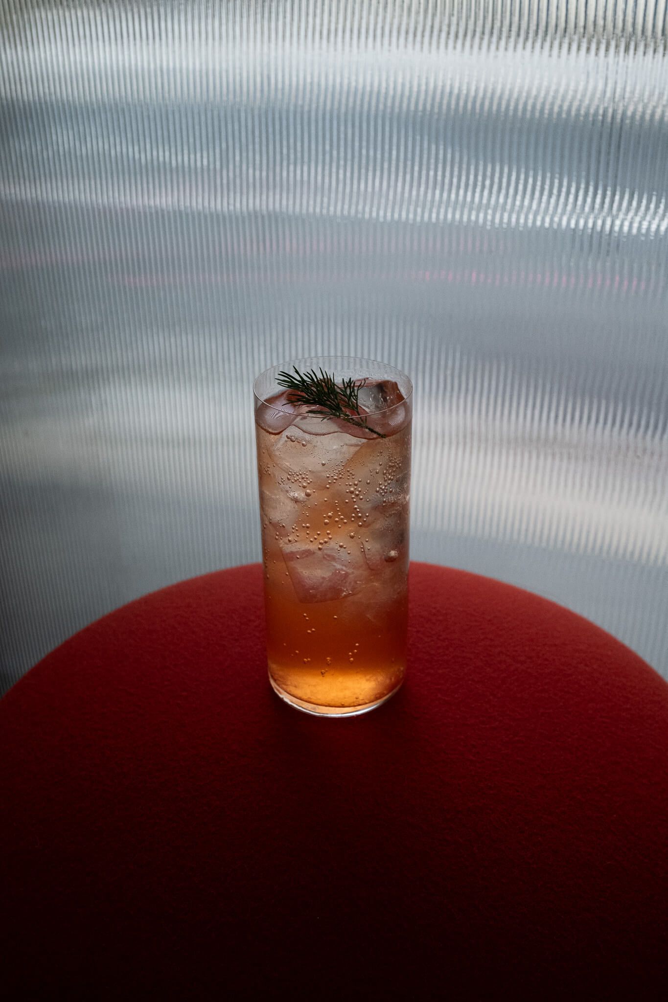 An orange highball cocktail with a rosemary garnish on a red table, backlit by a ribbed window.