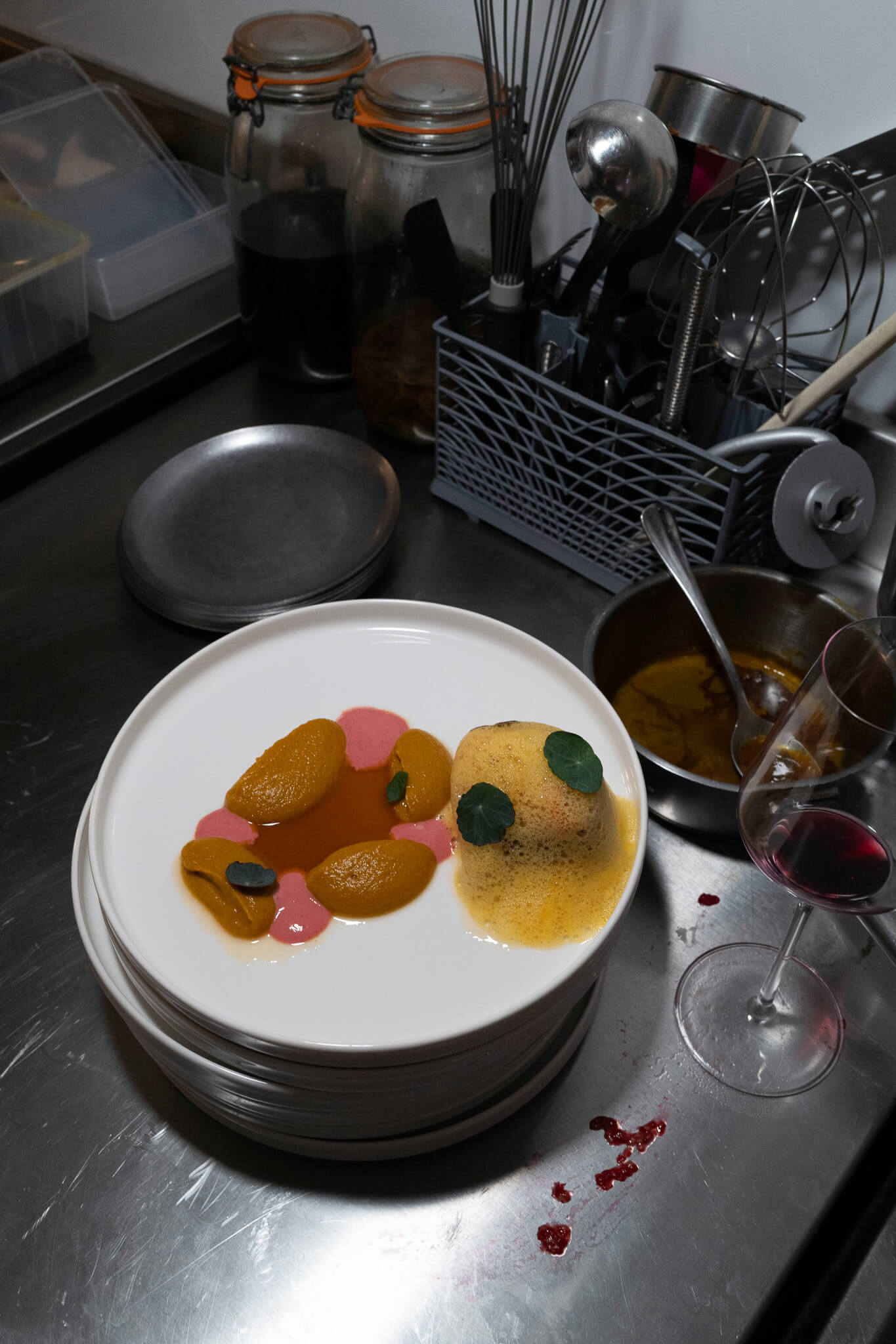 Overhead shot of a stacked plate presentation in a professional kitchen, with pink-sauced dumplings and greens beside a golden sauce, next to a glass of red wine on a steel counter.
