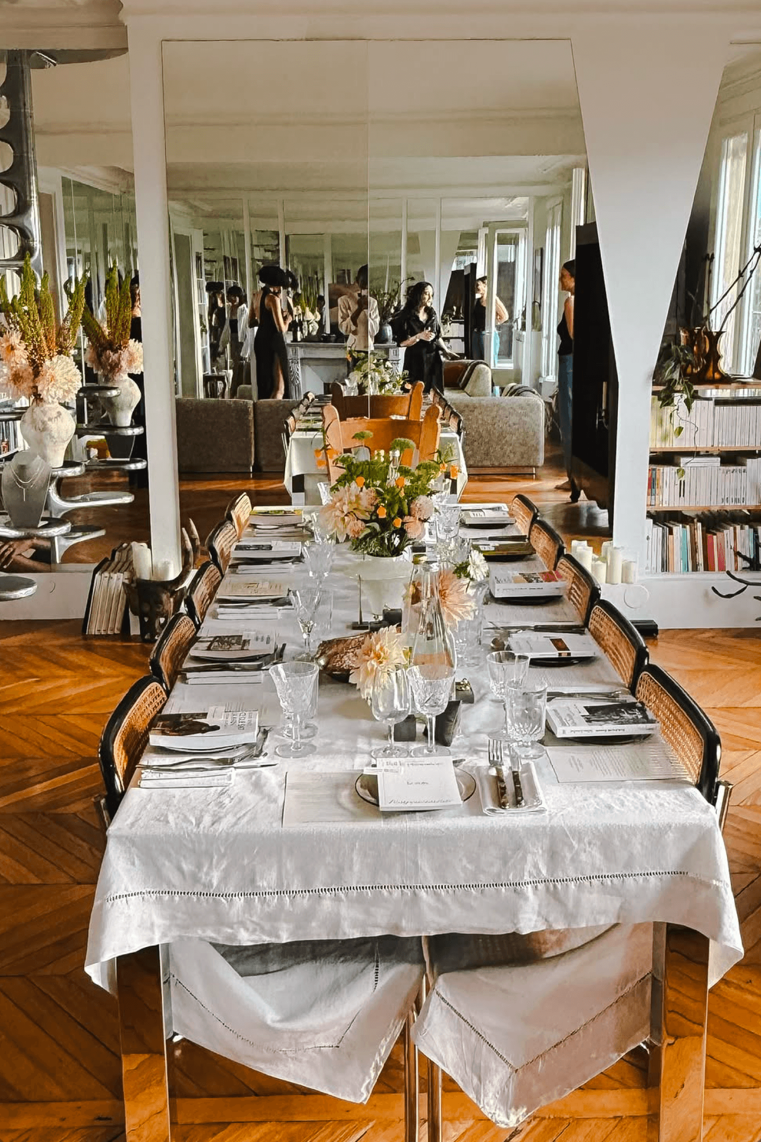 Wide shot of a long dinner table set in a mirrored, book-lined apartment with guests seated throughout.