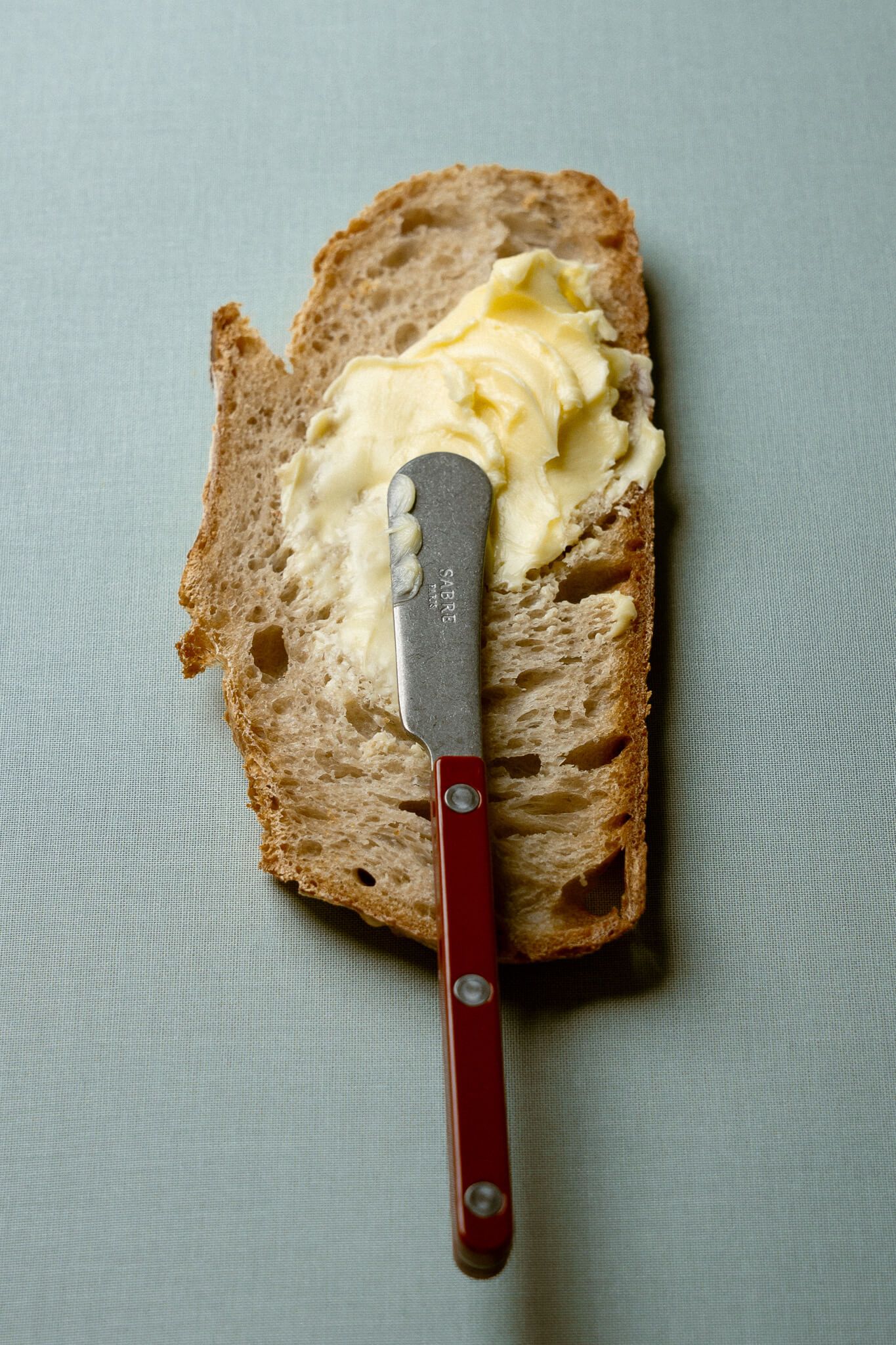 A silver butter knife with a red handle spreading butter on a slice of sourdough bread, on a light blue/grey background