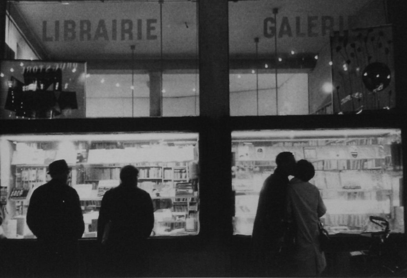 Black and white photograph of La Hune bookshop-gallery's window at night, with silhouettes of passersby