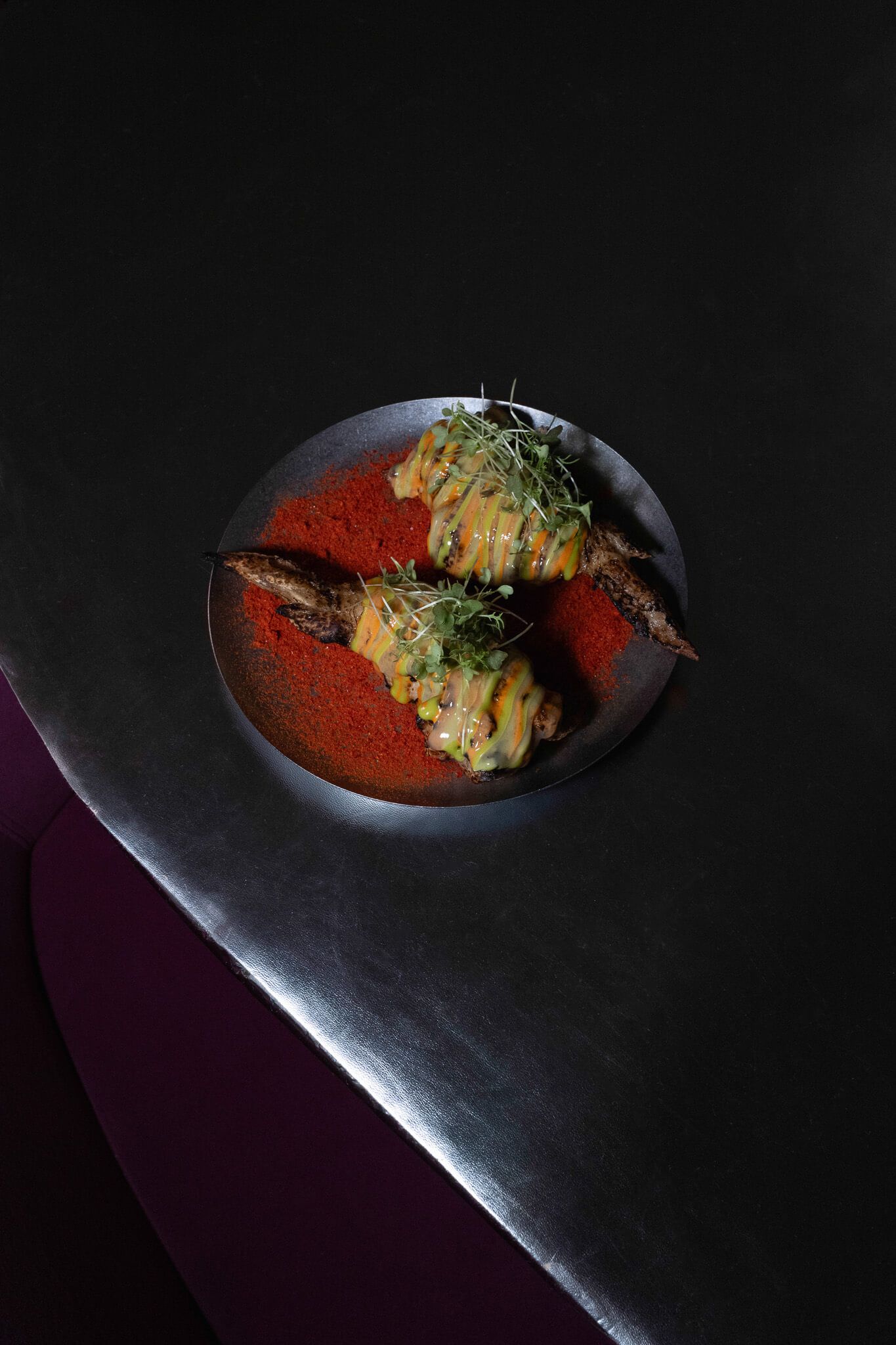 Overhead view of a plated dish with two chicken thighs, orange sauce drizzle, and micro greens on a dark red-orange spice bed, set on a silver table.