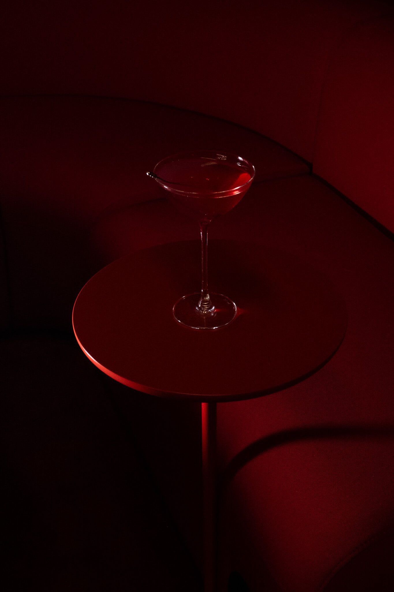 A red cocktail in a coupe glass on a glossy red table, lit dramatically against a dark red sofa.