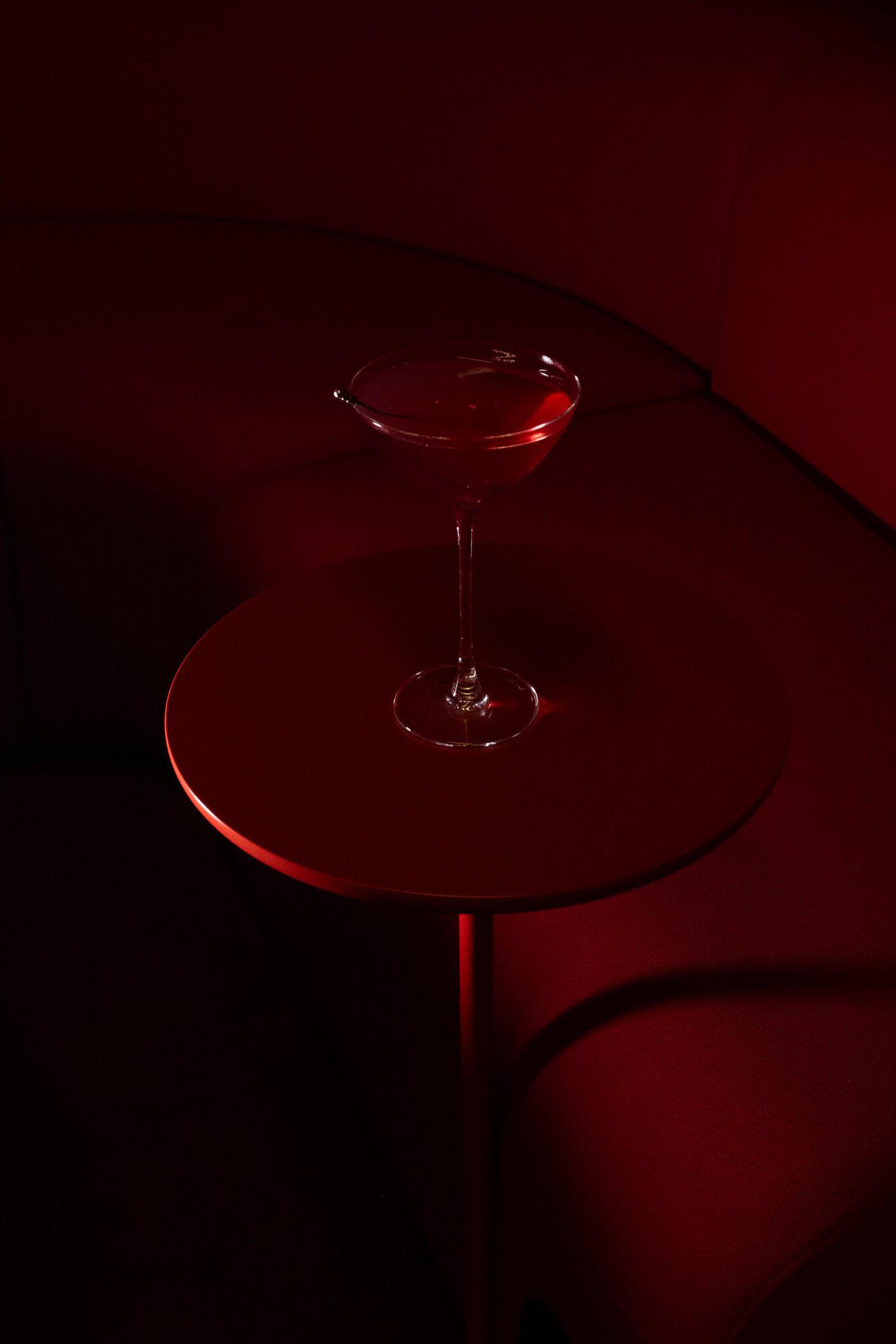 A red cocktail in a coupe glass on a glossy red table, lit dramatically against a dark red sofa.