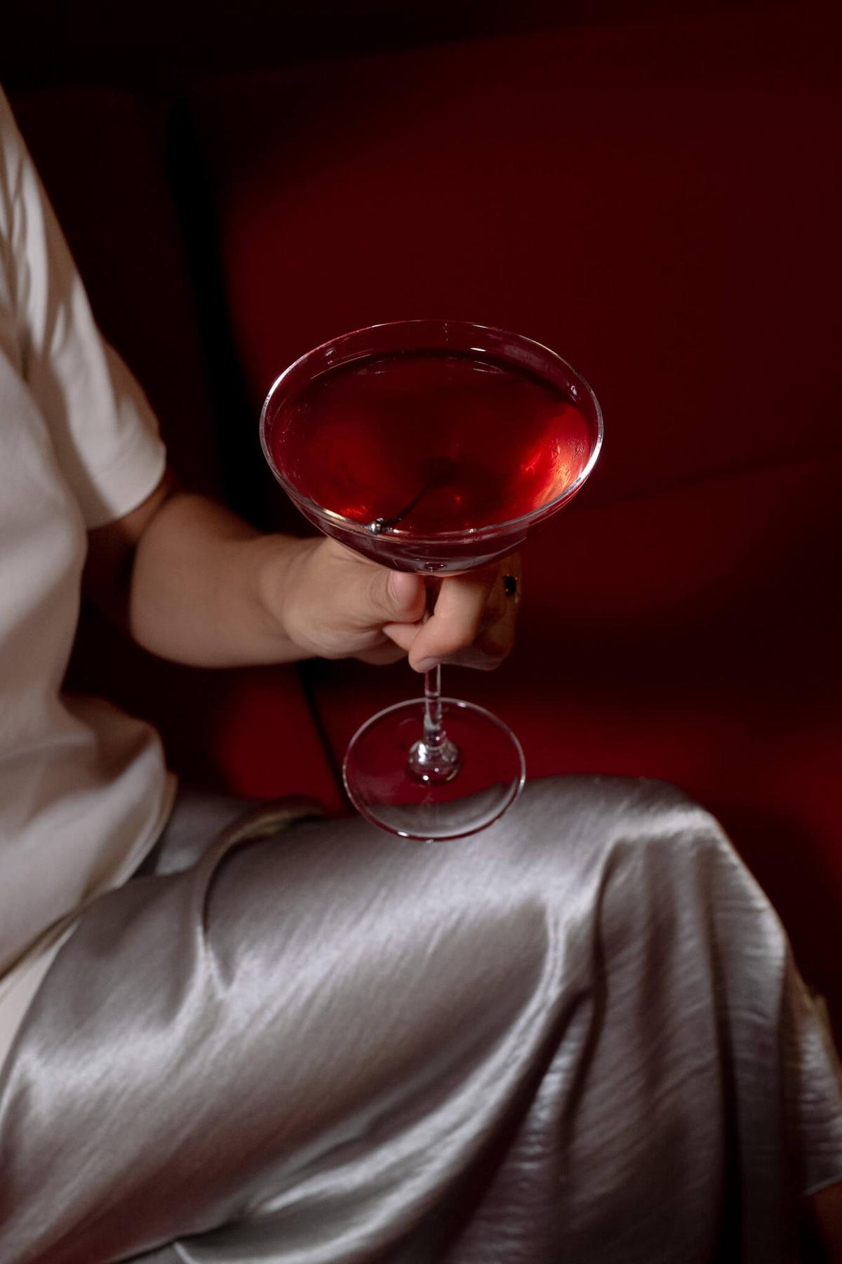 Close-up of a person in a silver satin outfit holding a red cocktail in a coupe glass, seated on a red velvet banquette.