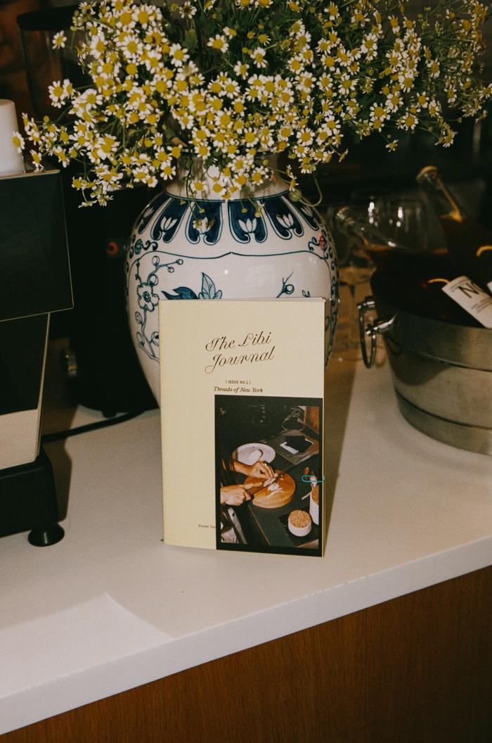 The journal displayed upright inside a blue-and-white patterned vase filled with daisies, on a white counter in a café setting.