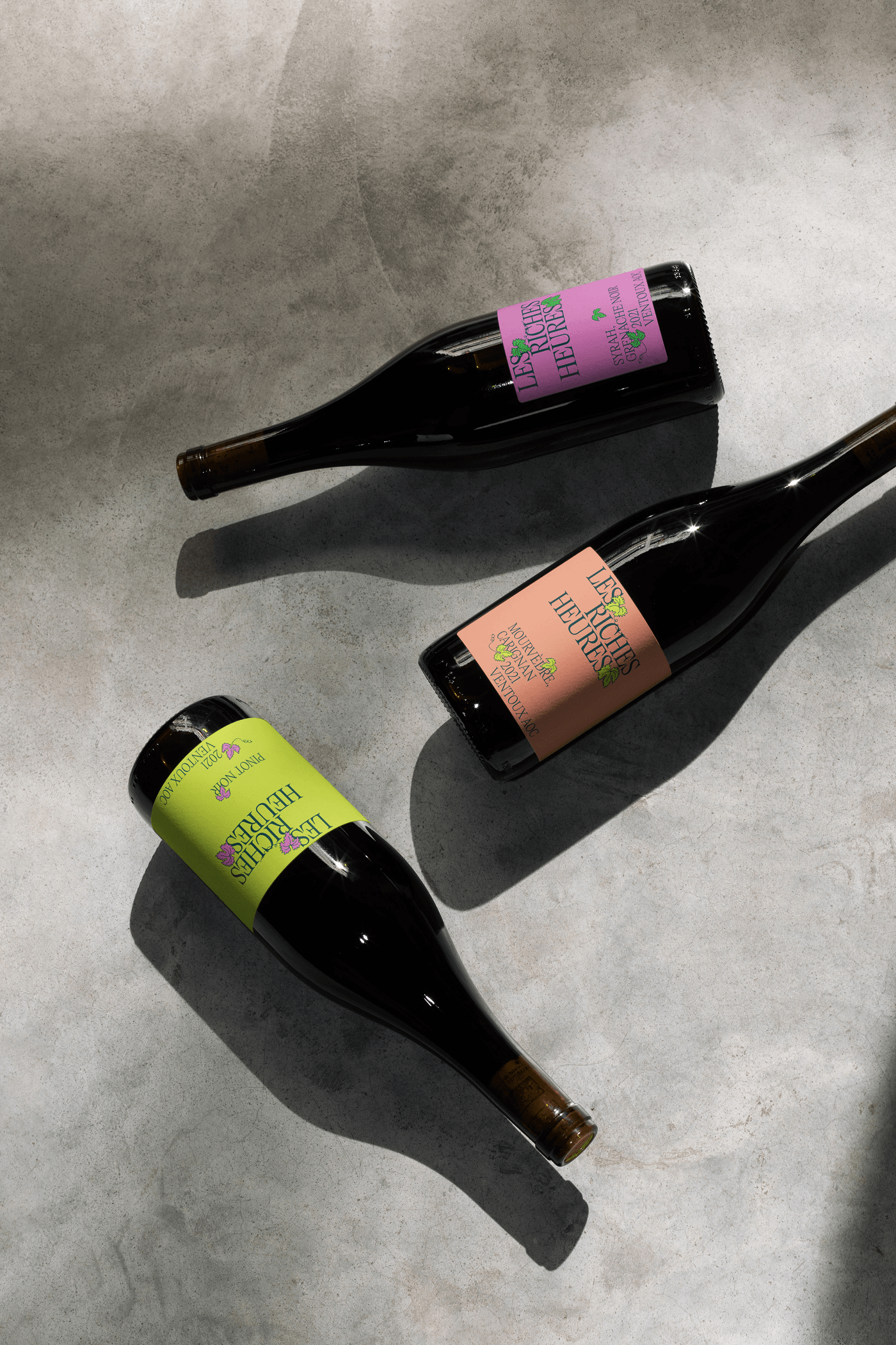 Overhead shot of three wine bottles with fuchsia, lime green, and salmon labels laid on a gray textured surface, shadows raking across.