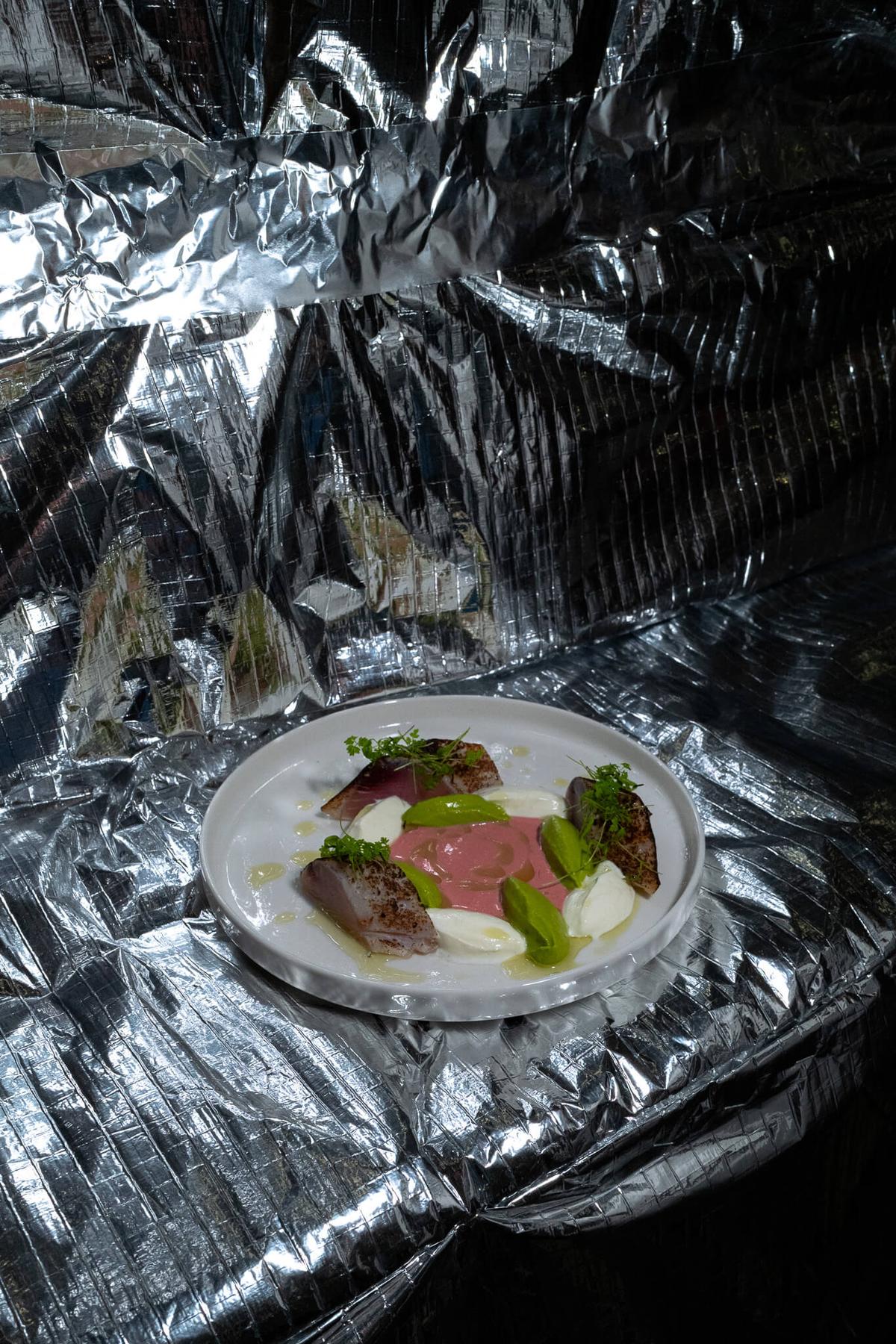 A plated dish of cured fish, greens, and sauce dots, photographed on crinkled silver foil.