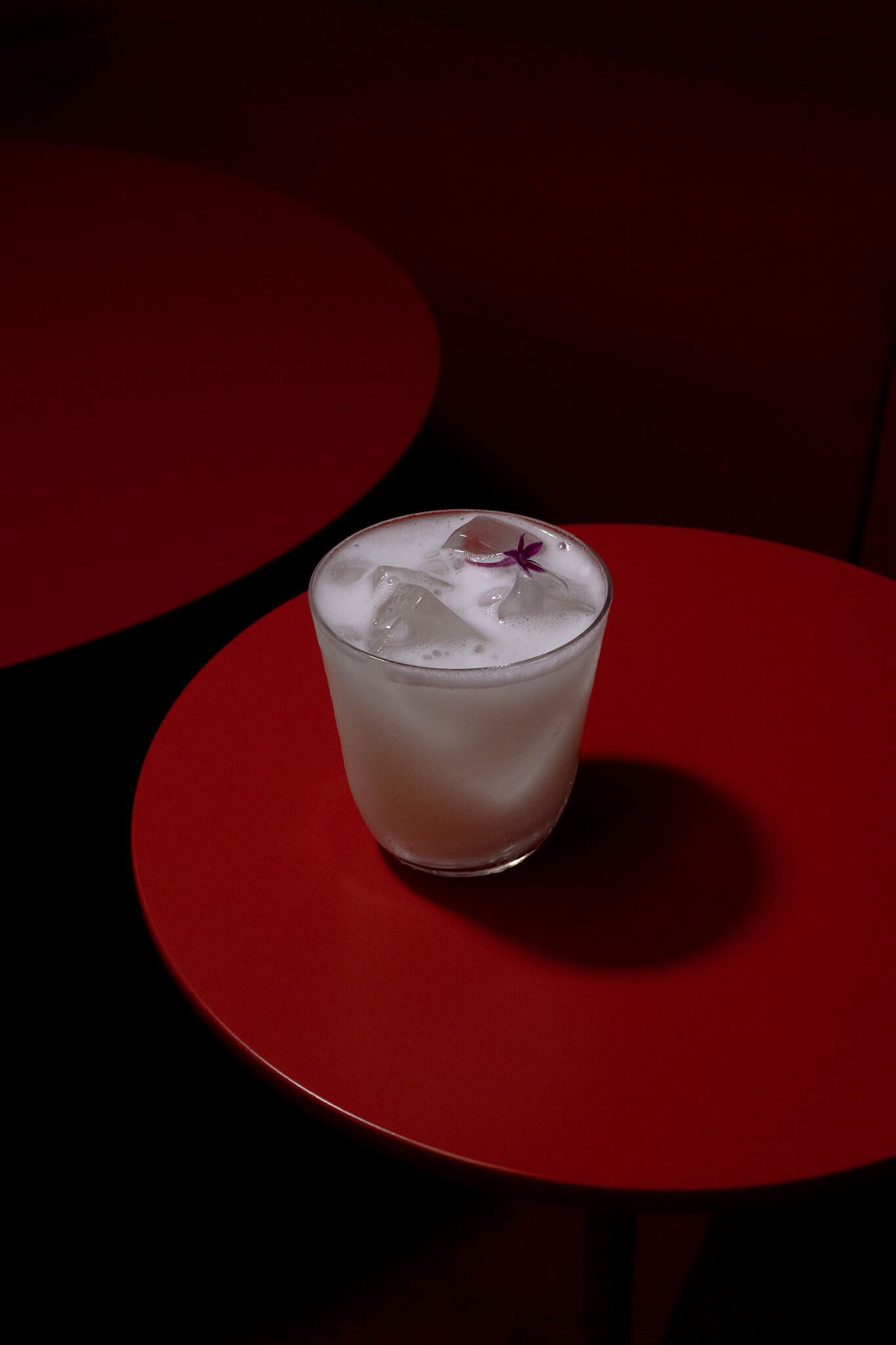 A cloudy white cocktail with ice and dried flower garnish, photographed on a red side table against a dark red backdrop.