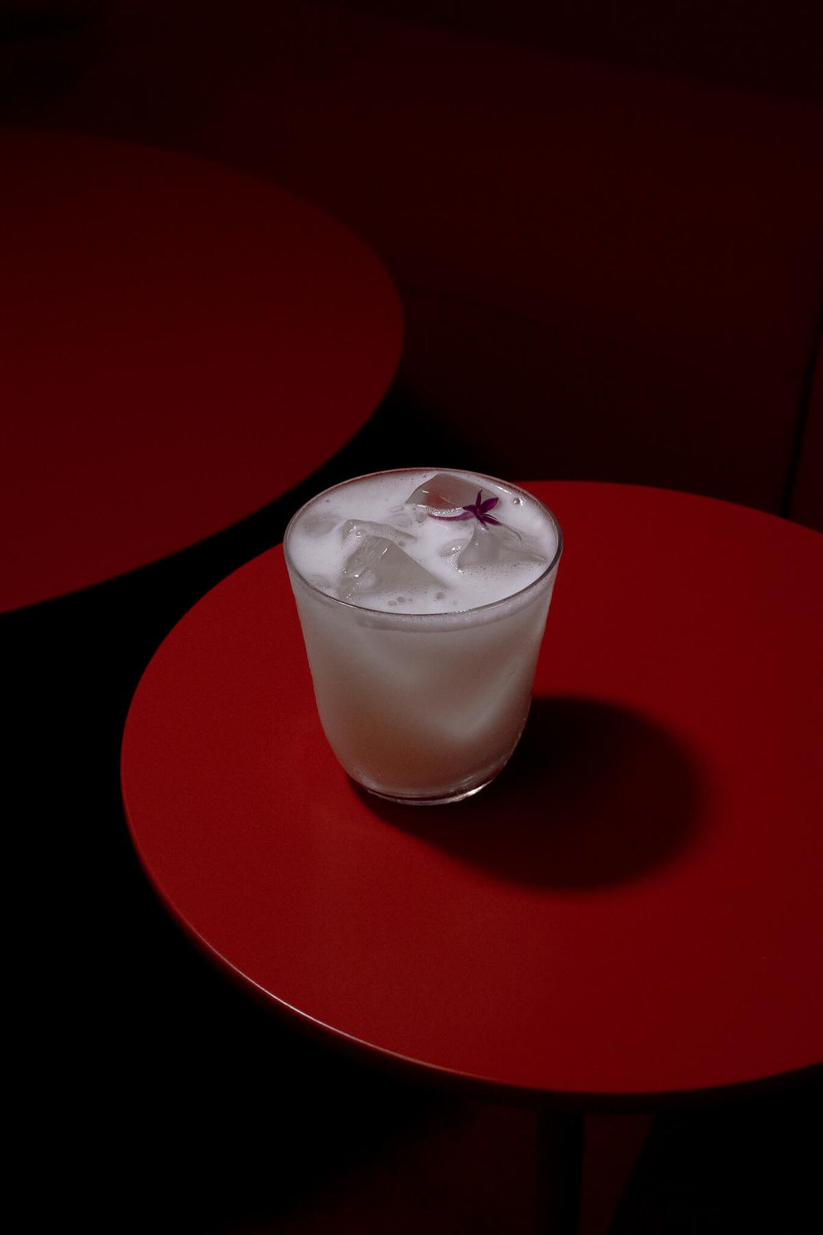 A cloudy white cocktail with ice and dried flower garnish, photographed on a red side table against a dark red backdrop.