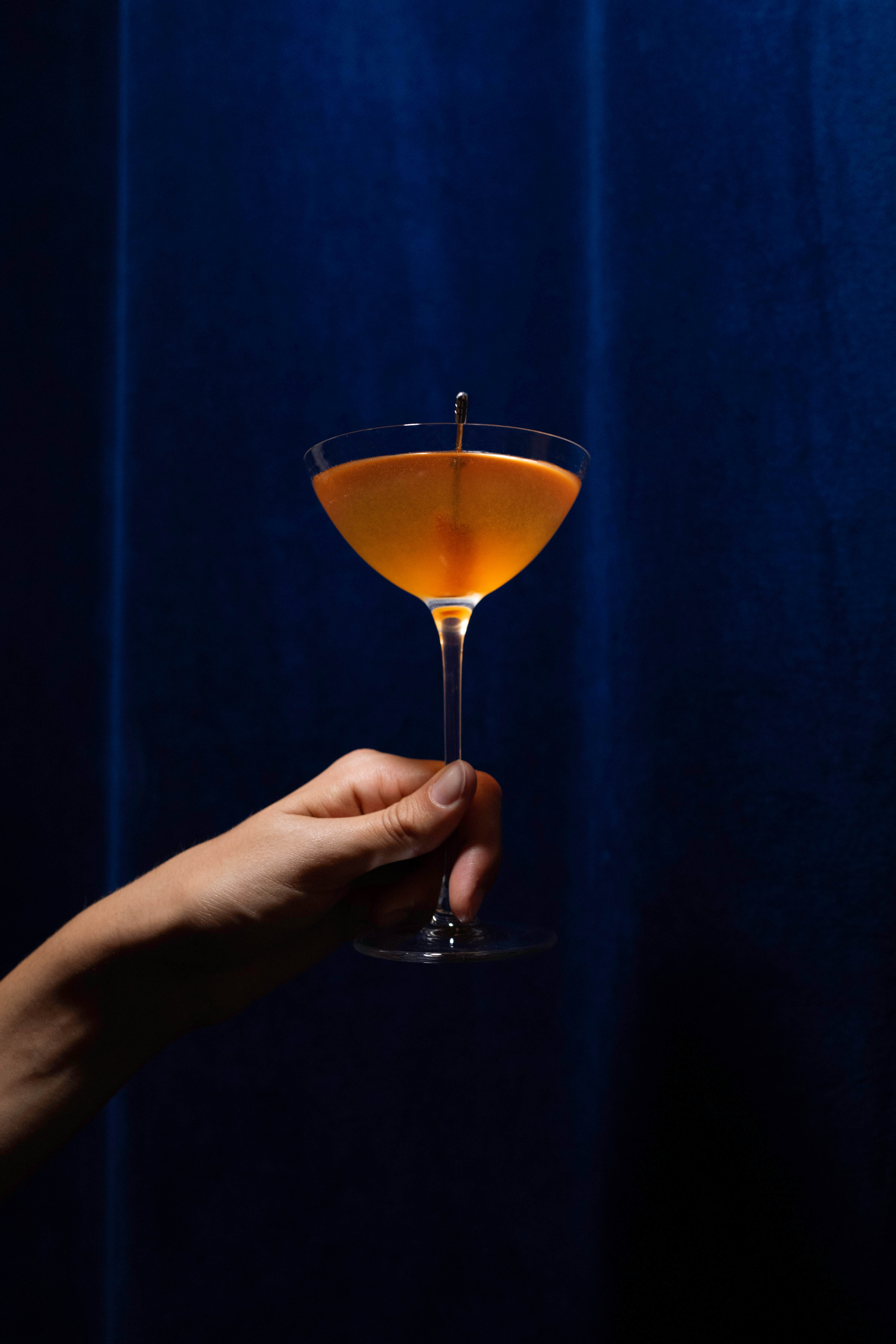 A hand holding an orange cocktail in a coupe glass against a deep blue velvet curtain backdrop.