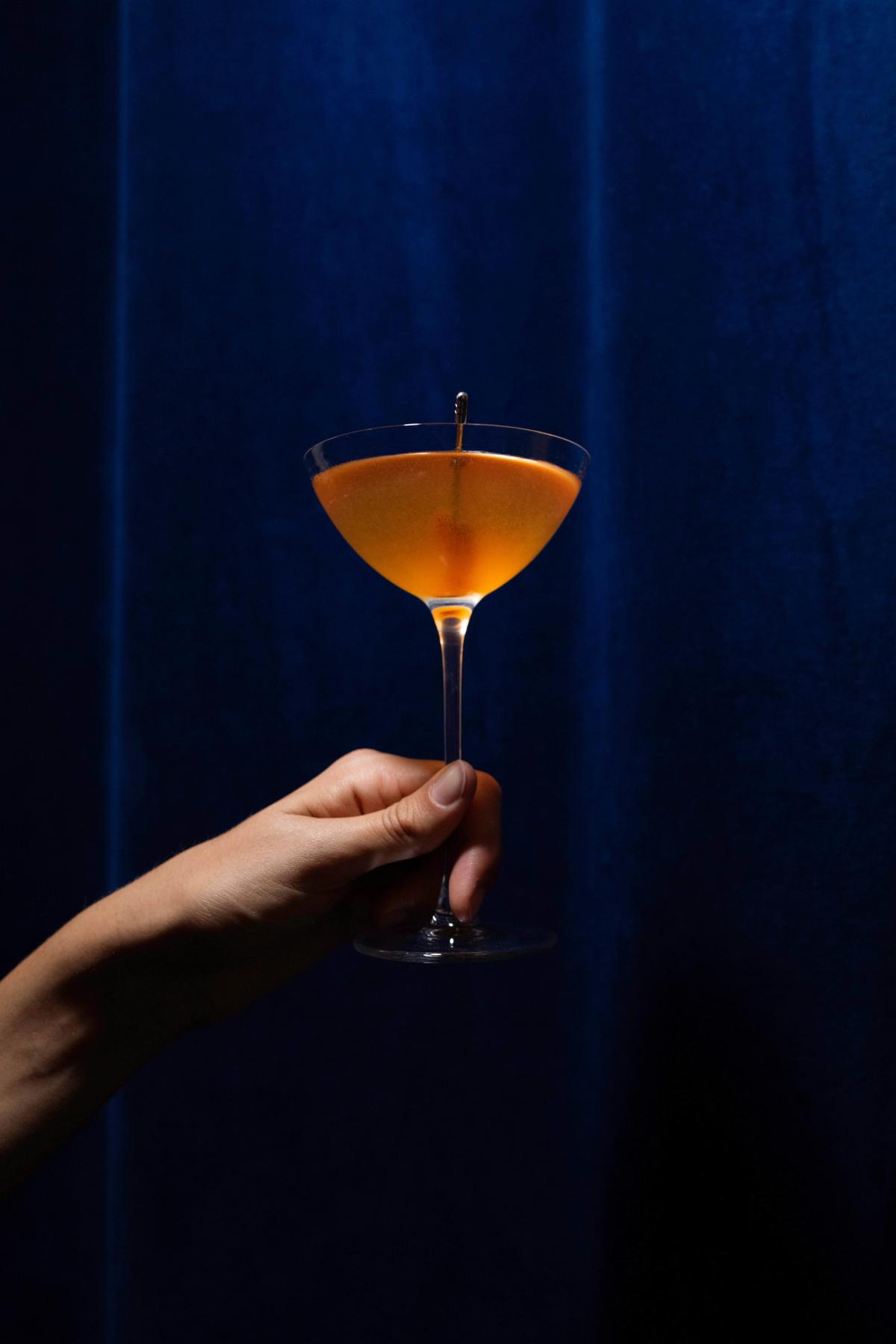 A hand holding an orange cocktail in a coupe glass against a deep blue velvet curtain backdrop.