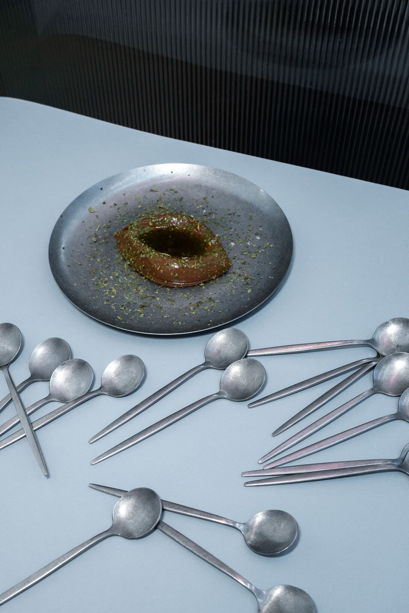 Overhead shot of a dark chocolate ganache dessert covered in olive oil and dusted with herbs on a metal plate, surrounded by a scattered arrangement of small spoons on a pale blue surface.