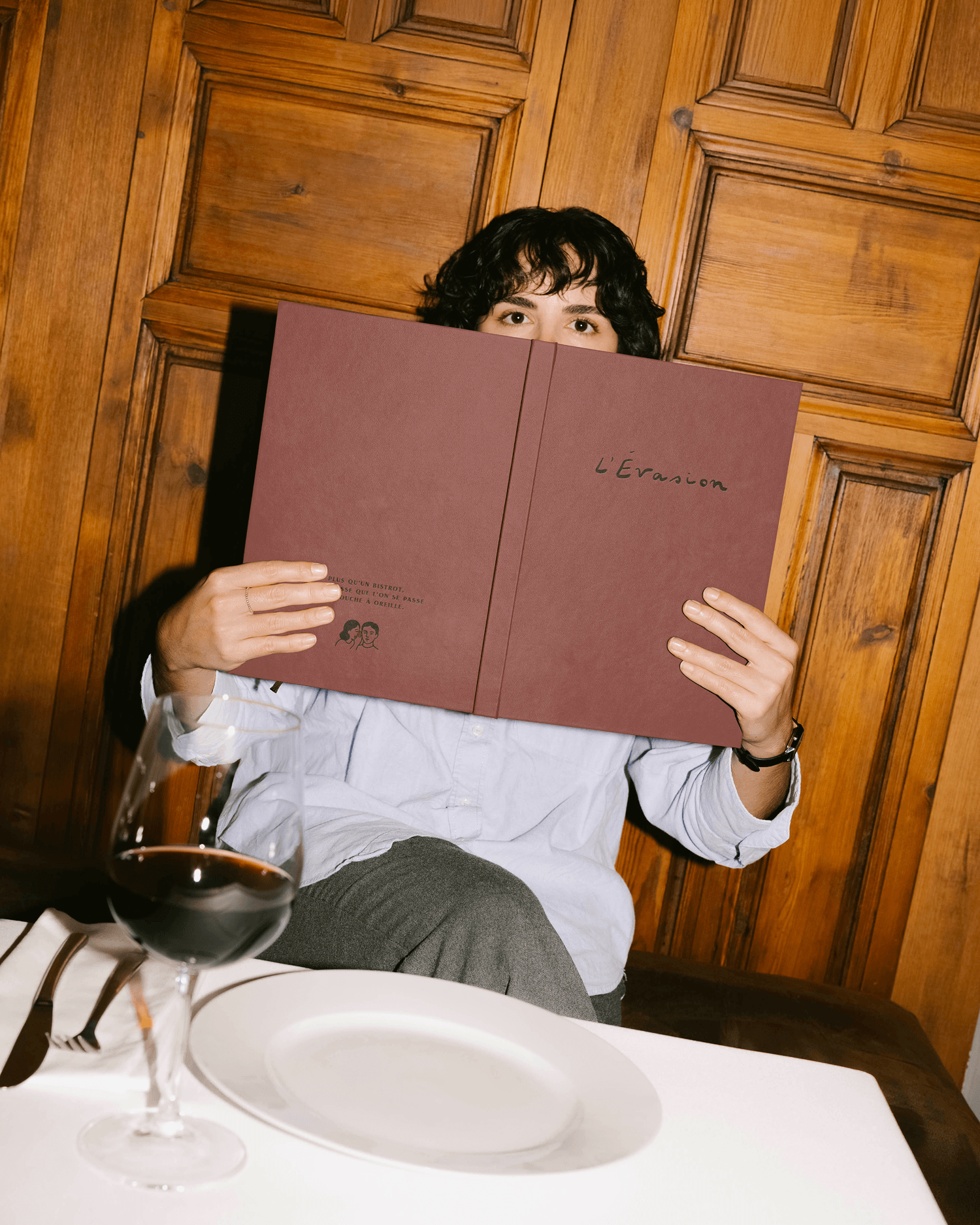 A person peeking over an open burgundy menu in front of wood-paneled wall, wine glass and plate visible below.