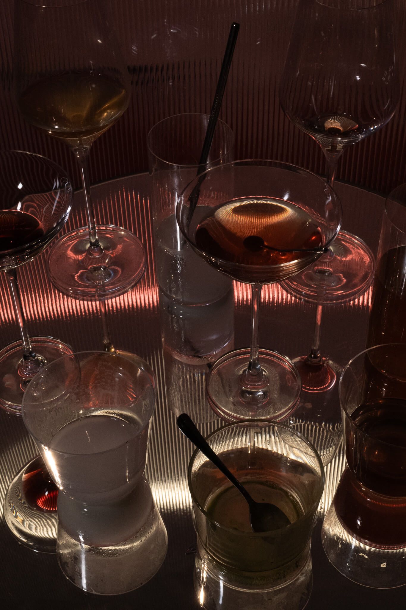 An assortment of cocktail glasses catching pink light on a reflective table.