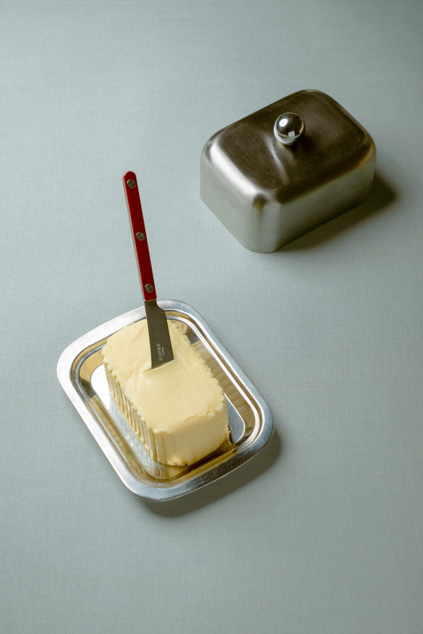 A mutt of butter on a silver dish, with a silver butter knife with a red handle planted in it, on a light blue/grey background