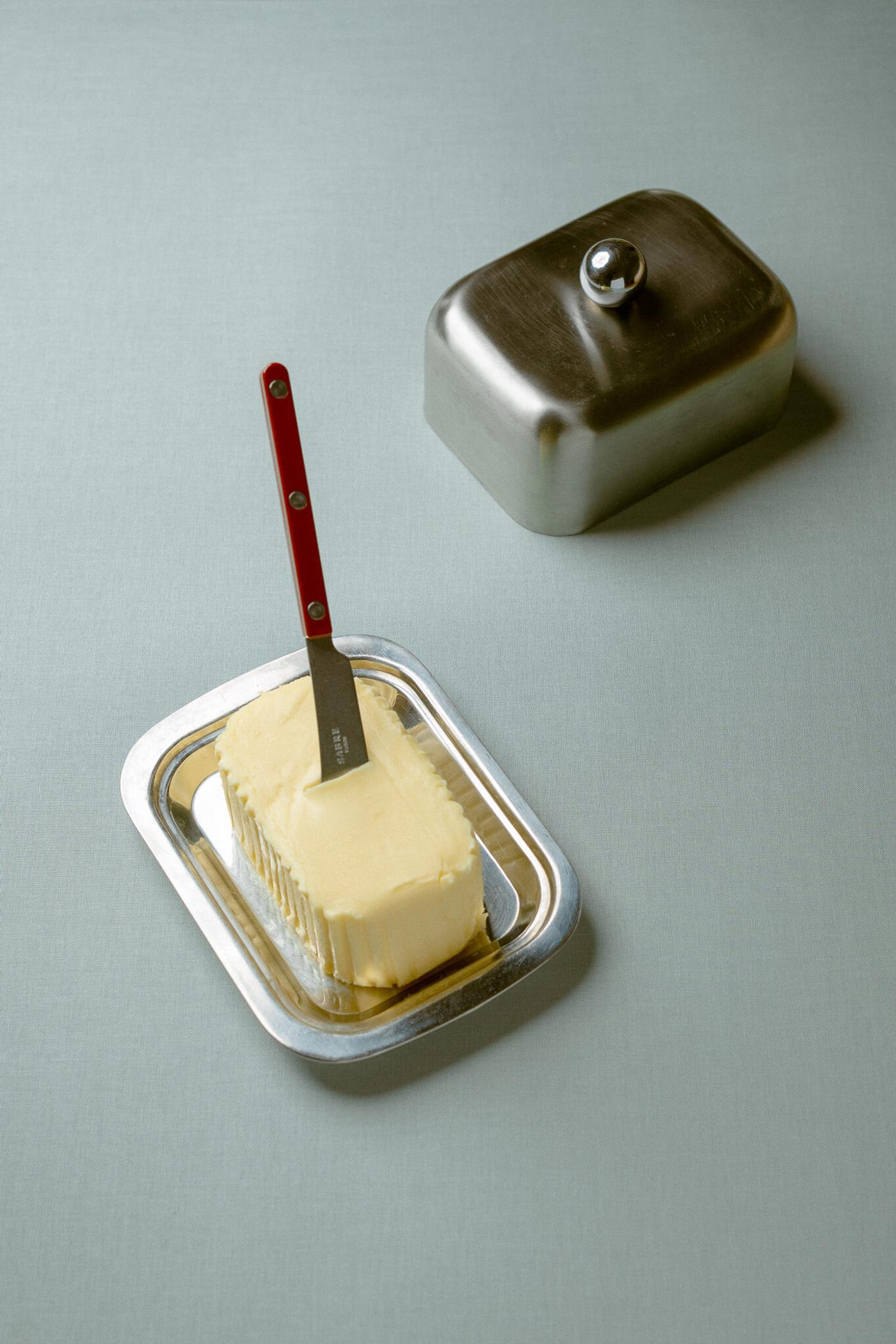 A mutt of butter on a silver dish, with a silver butter knife with a red handle planted in it, on a light blue/grey background