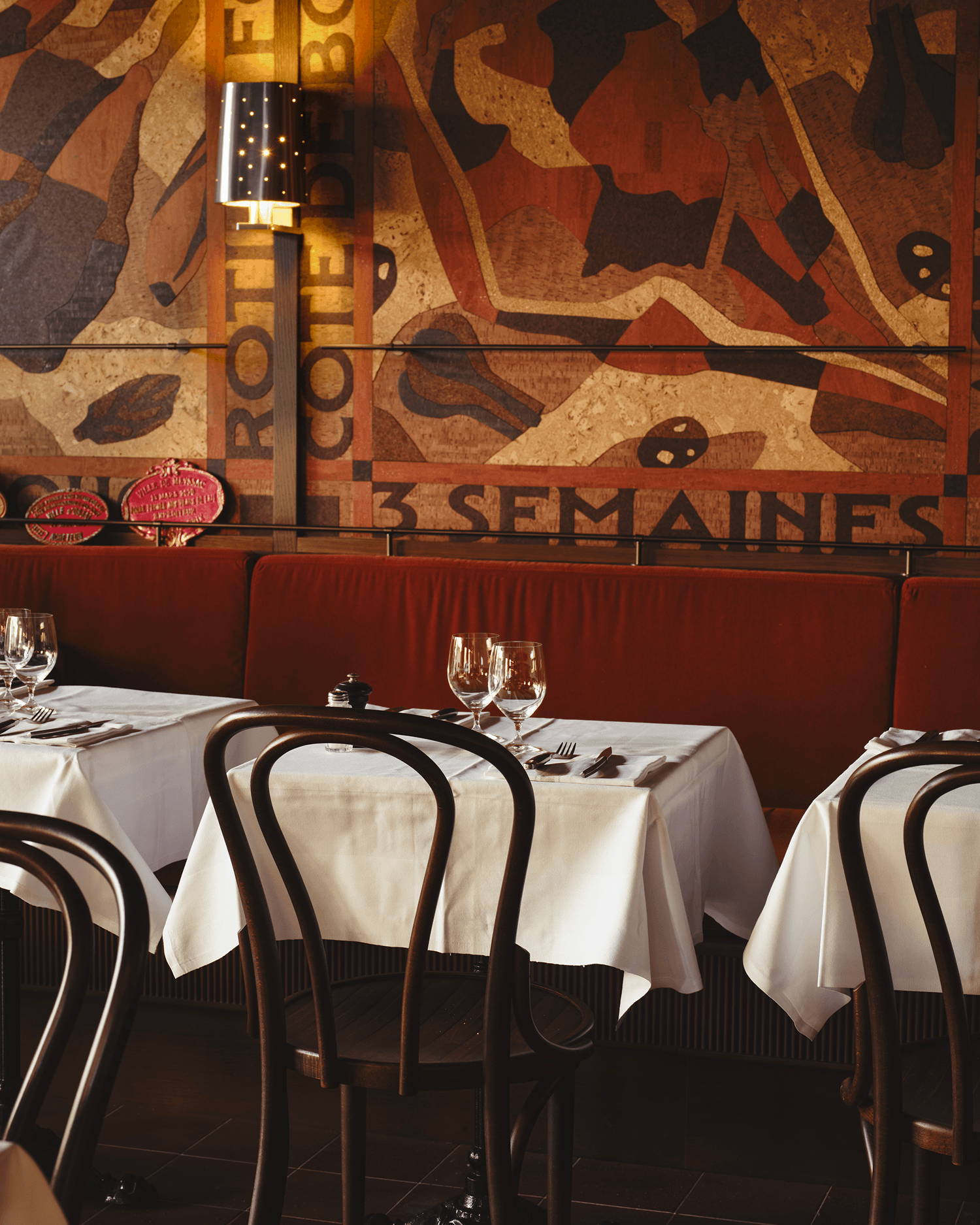 Restaurant dining room interior with white tablecloths, dark bentwood chairs, and a patterned cork mural.