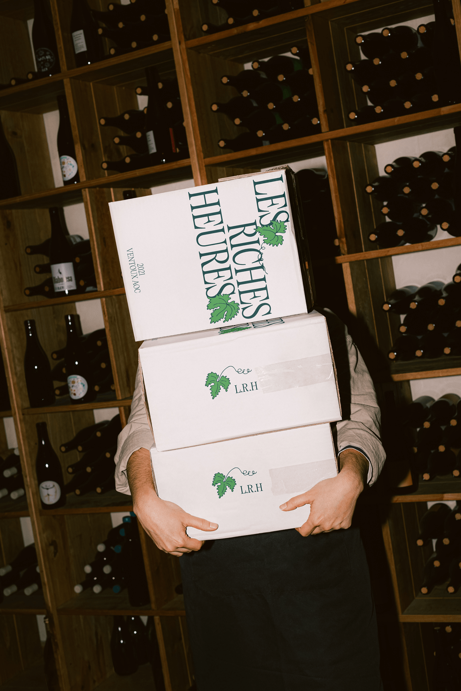 A person carrying stacked "Les Riches Heures" shipping boxes through a wine cellar lined with bottle racks.