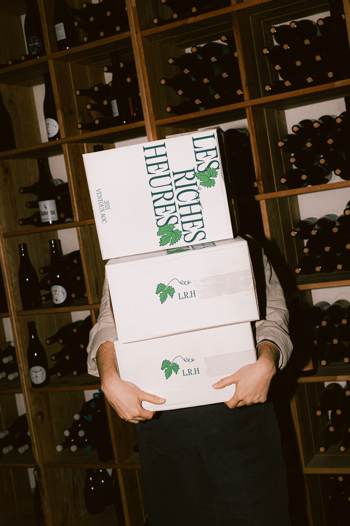 A person carrying stacked "Les Riches Heures" shipping boxes through a wine cellar lined with bottle racks.
