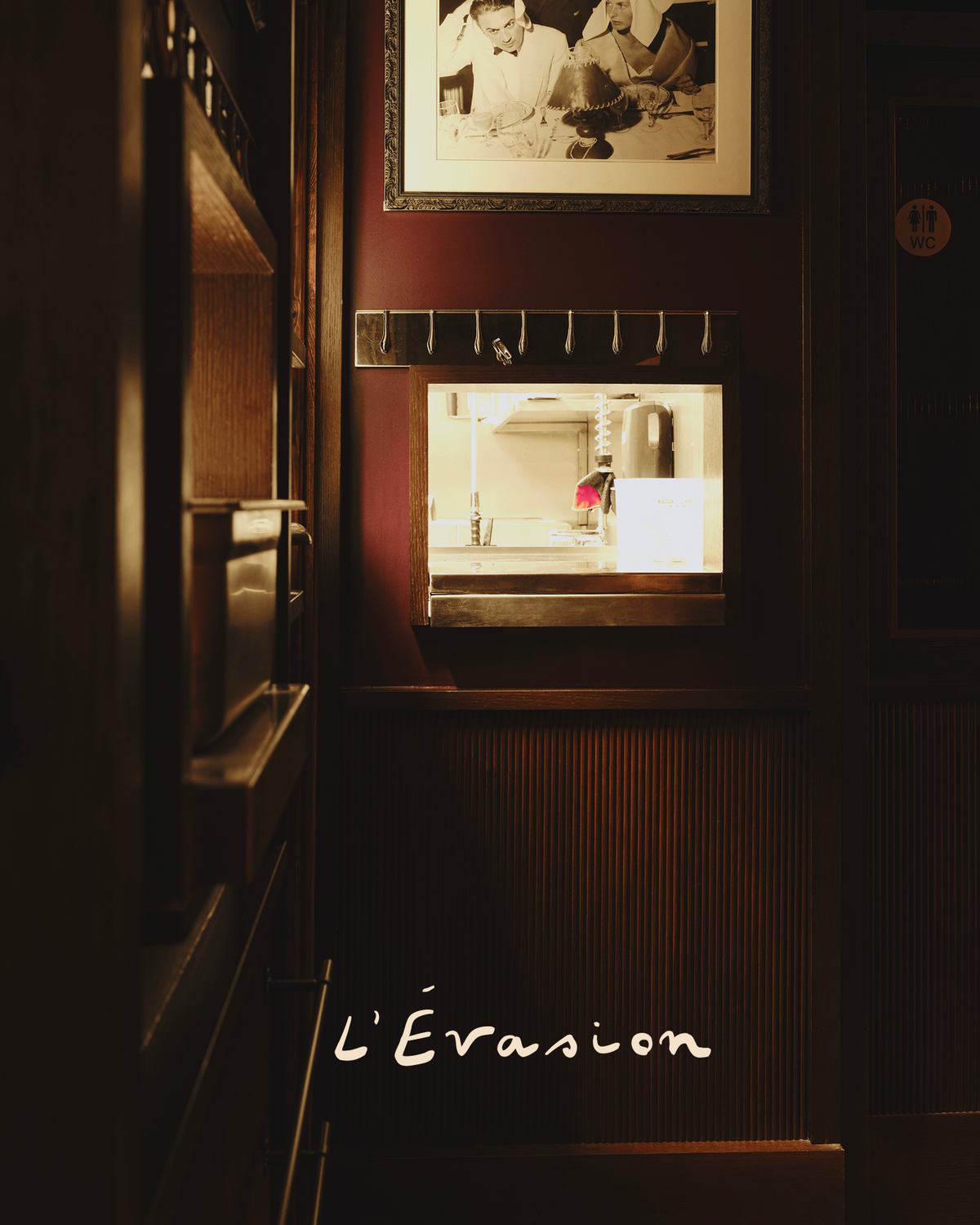 Dim restaurant interior detail showing a vintage photo, shelving, and warm pendant lighting, with the "L'Évasion" logo overlaid.