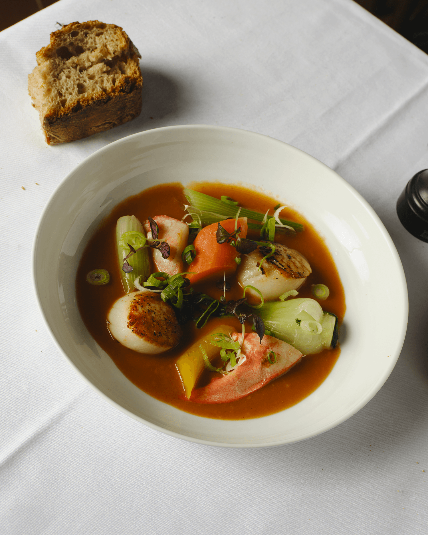 Overhead shot of a scallop and vegetable dish in broth, served with a torn piece of bread, on a white tablecloth.