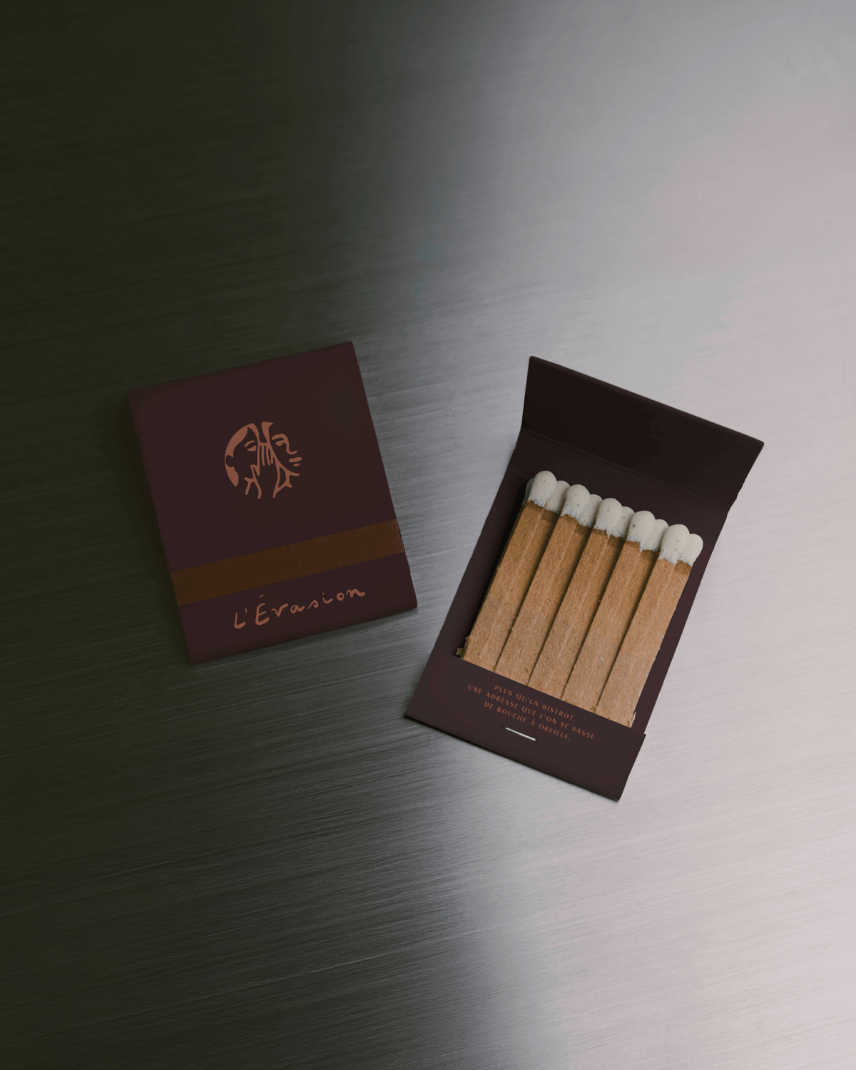 A burgundy matchbox printed with the L'Évasion logo, opened to show wooden matches, photographed on a brushed metal surface.