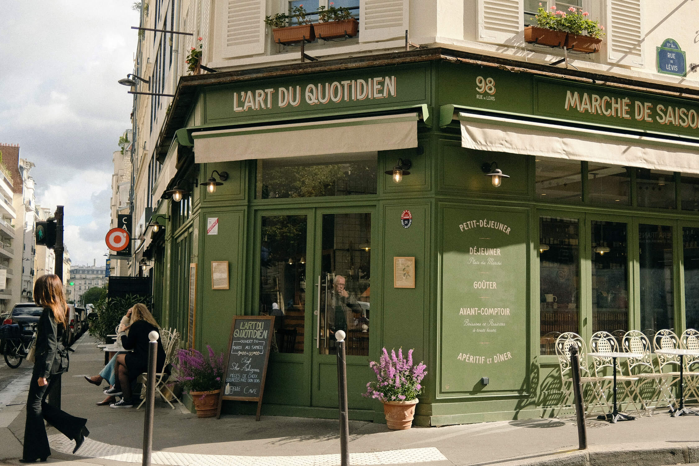 Street-level view of the restaurant's corner facade in daylight, showing the full signage, awnings, potted flowers, and passersby.