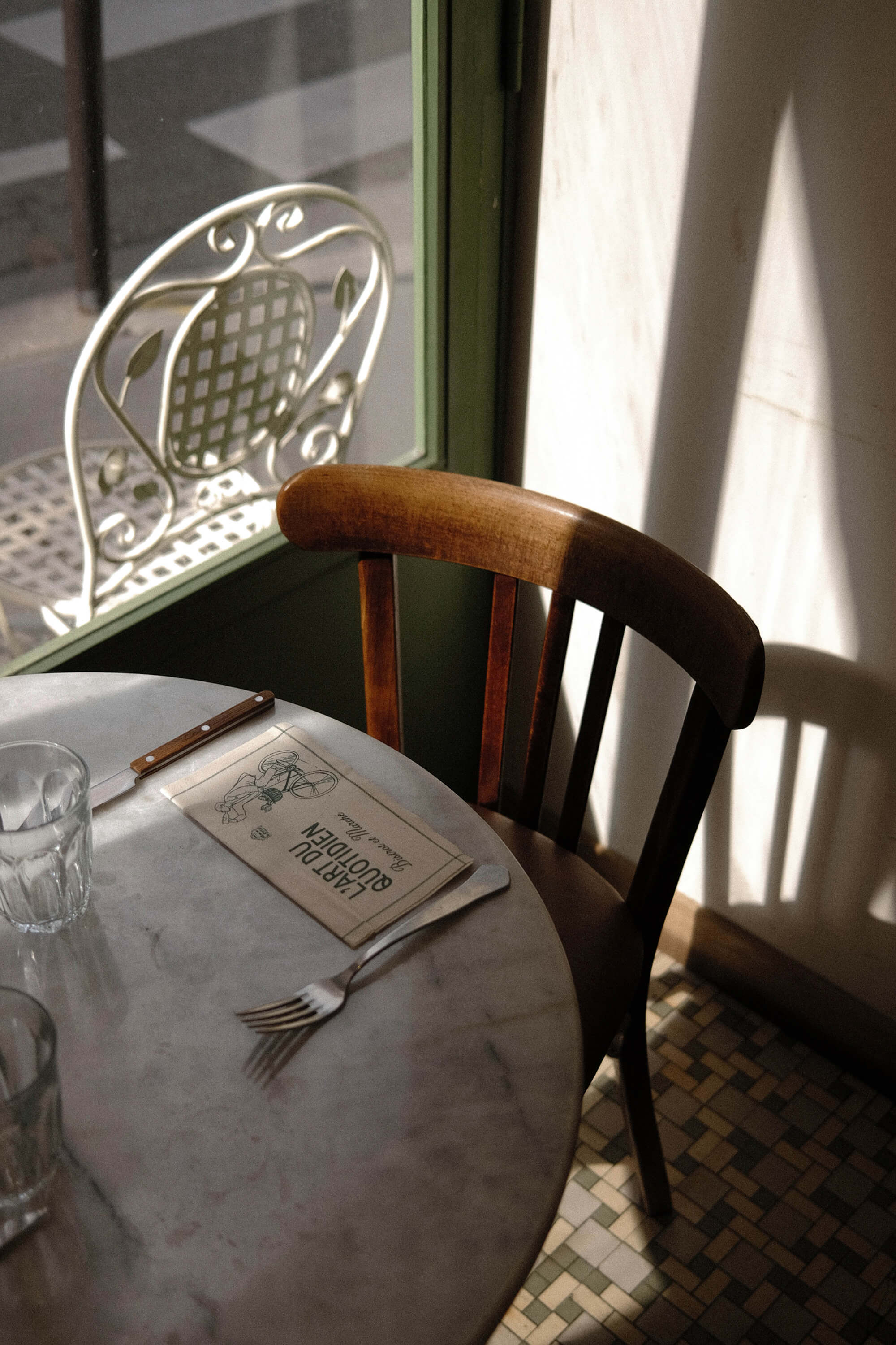 Close-up of a wooden bistro chair and marble table with a folded menu, lit by afternoon sun through the window.