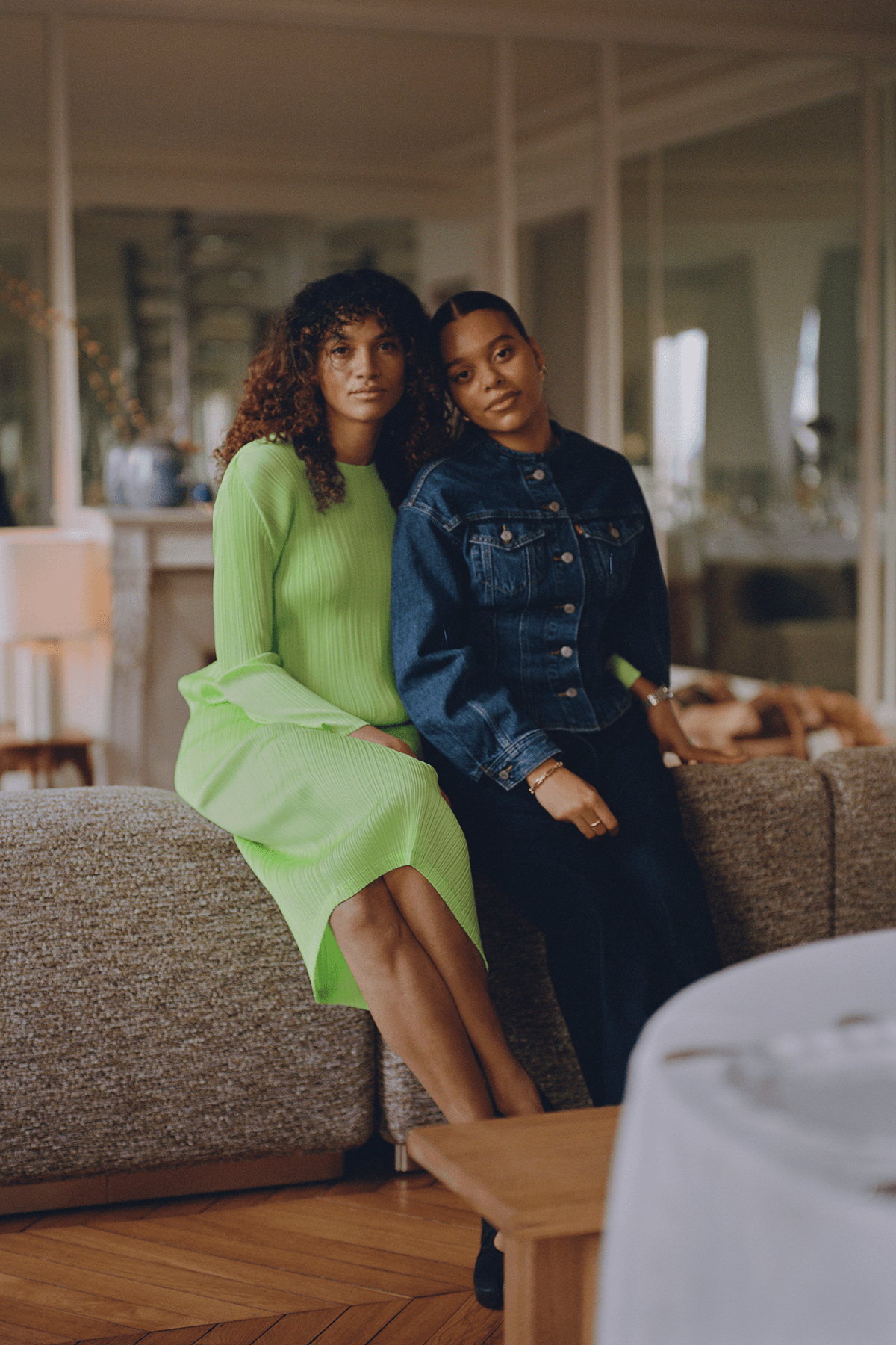 Laurianne Melierre and Romane Limone posing together on a sofa, one in a green knit dress and one in a denim jacket, in a softly lit Parisian apartment.