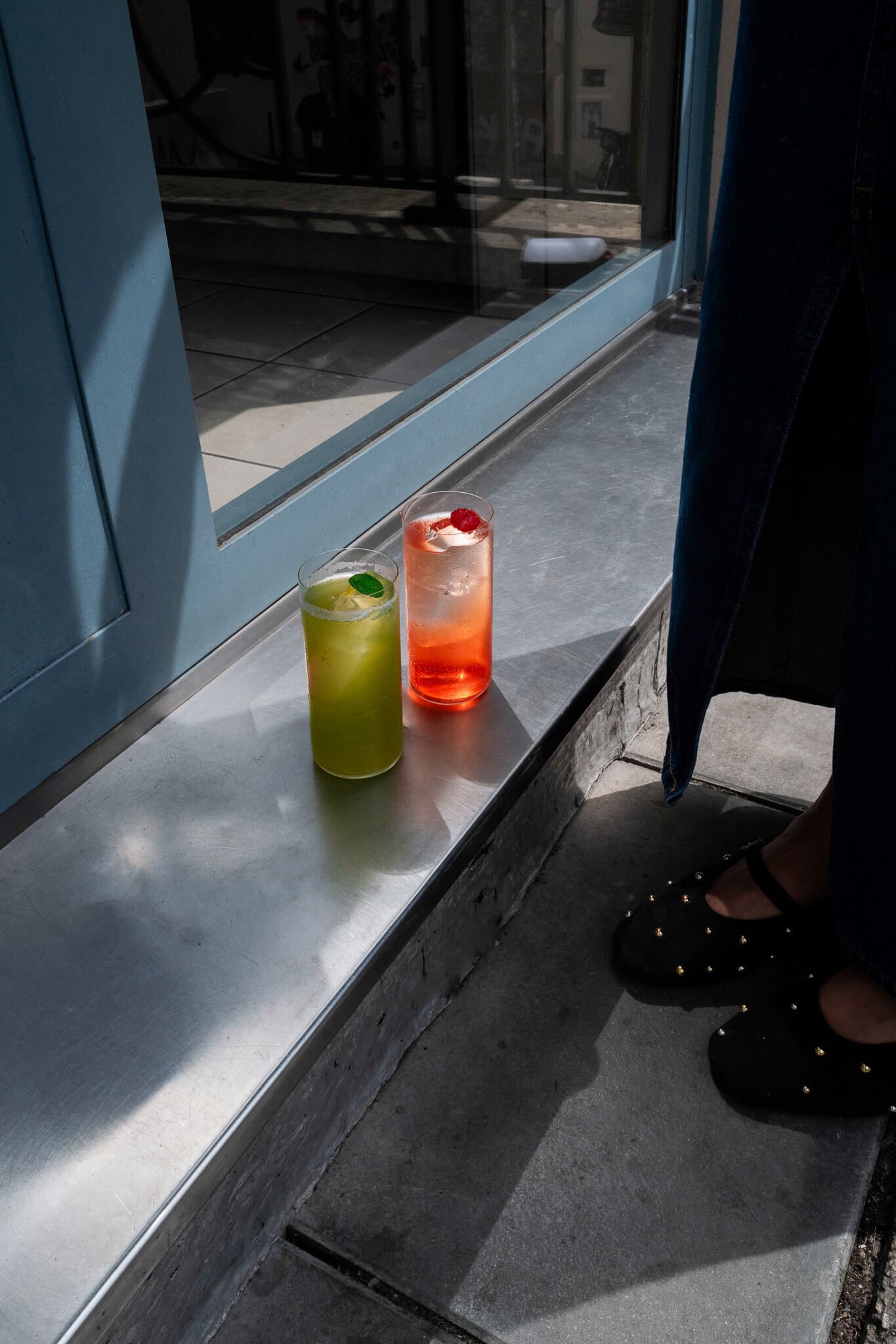 Two cocktails, one green and one pink, resting on a metal ledge by a window, with a person's feet visible below.