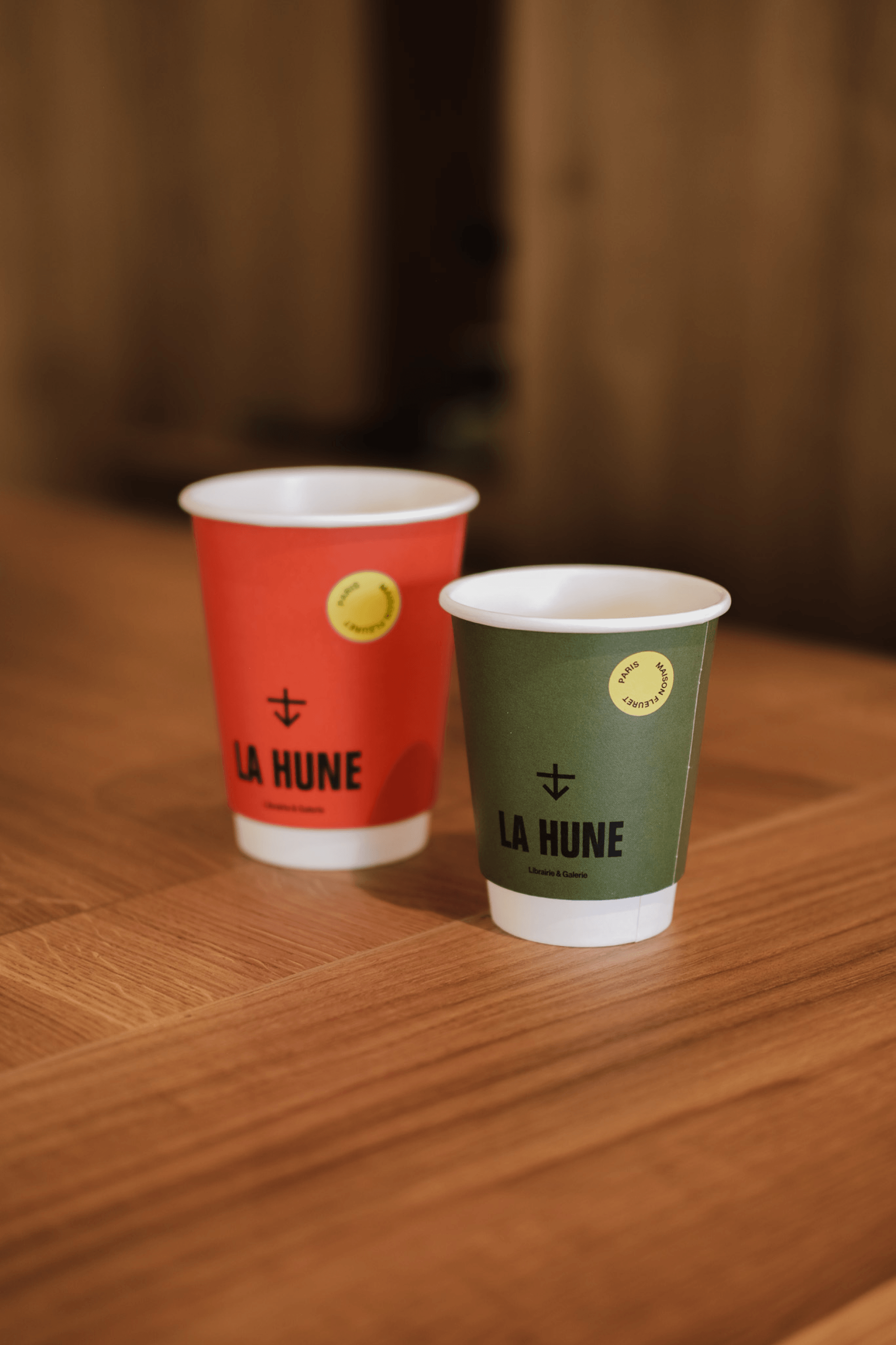 Two La Hune takeaway coffee cups in red and green, placed on a wooden bar.