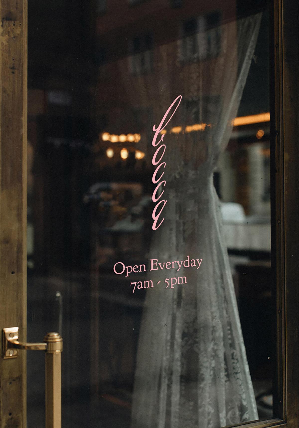 Close-up of the café's glass door at night, showing the pink cursive "Bocca" logo and "Open Everyday 7am - 5pm" text etched on the window, with warm interior lighting visible through sheer curtains.