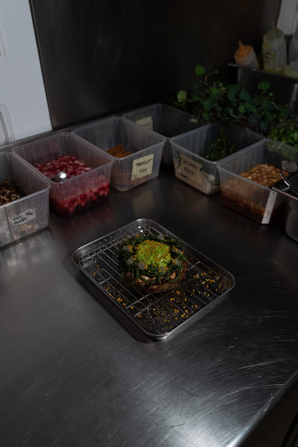 Overhead view of prep ingredients in small kitchen containers, including herbs, diced garnishes, and a small dish, on a steel counter.