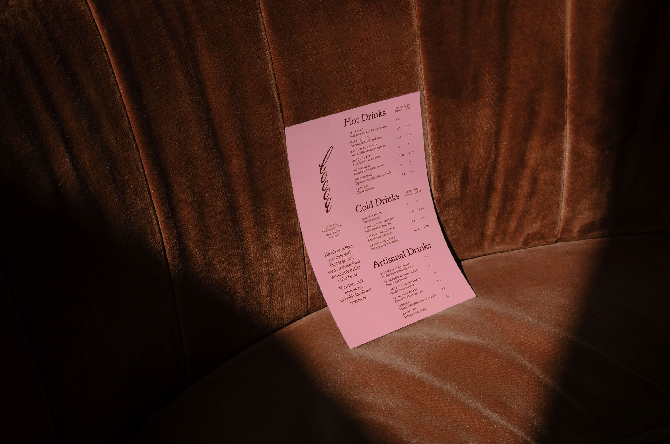 A pink drinks menu propped upright on a brown velvet banquette seat, lit by warm side lighting.