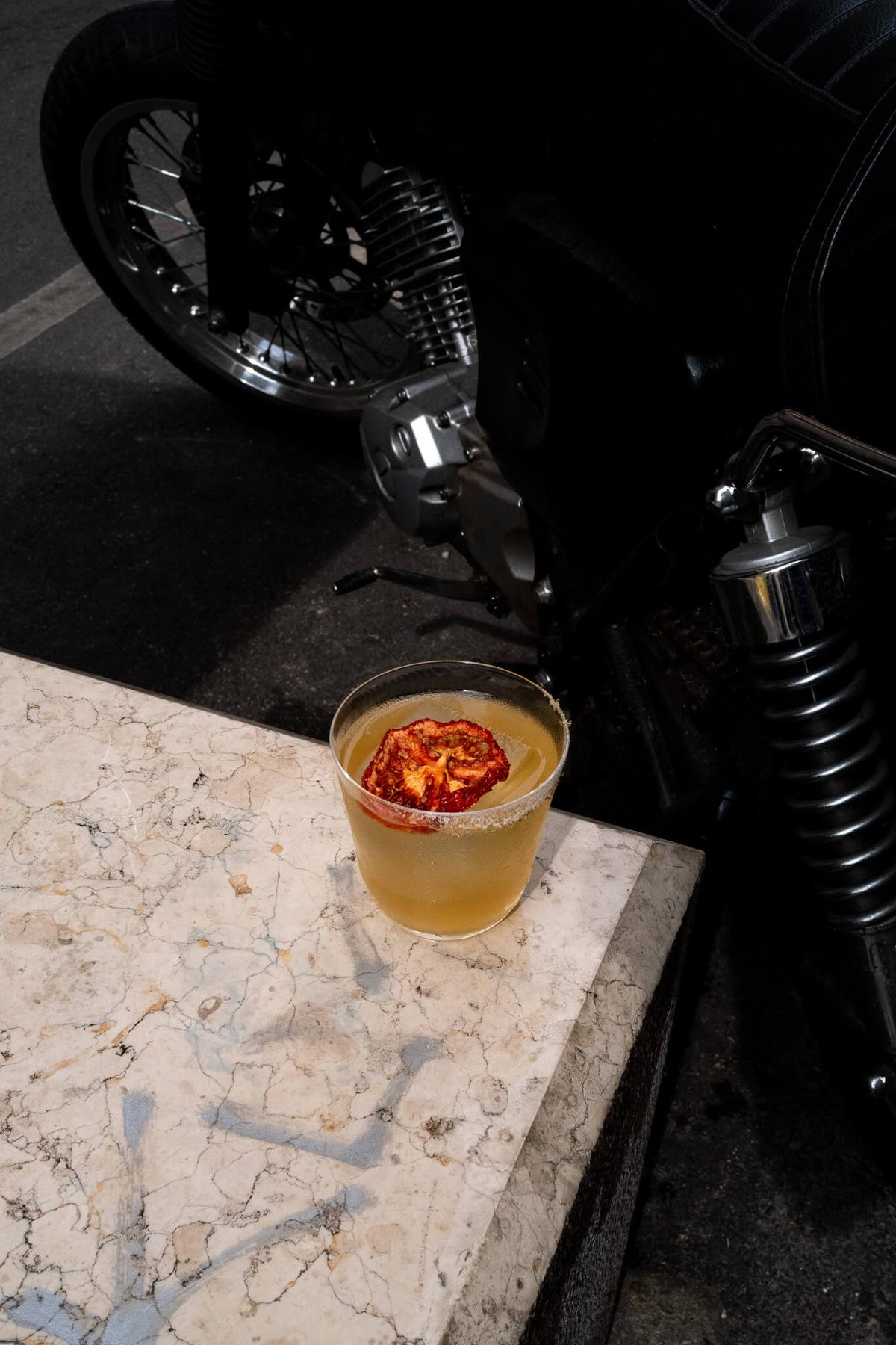 An amber cocktail with dried tomate garnish resting on a marble ledge, with a motorcycle parked in the dark background.
