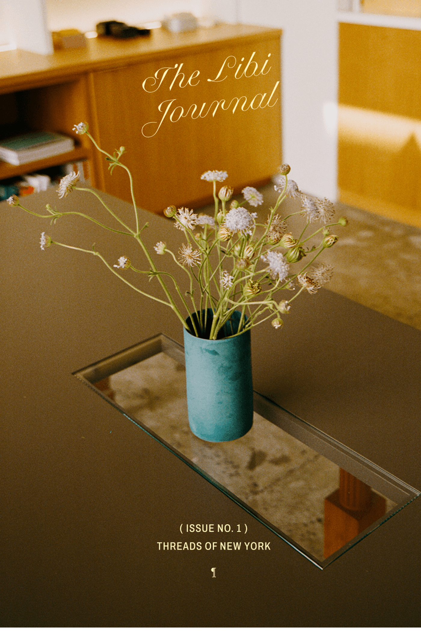 A copy of "The Libi Journal, Issue No. 1: Threads of New York" lying on a dark wood table next to a teal vase filled with wild flowers, styled on a mirrored tray in warm ambient light.