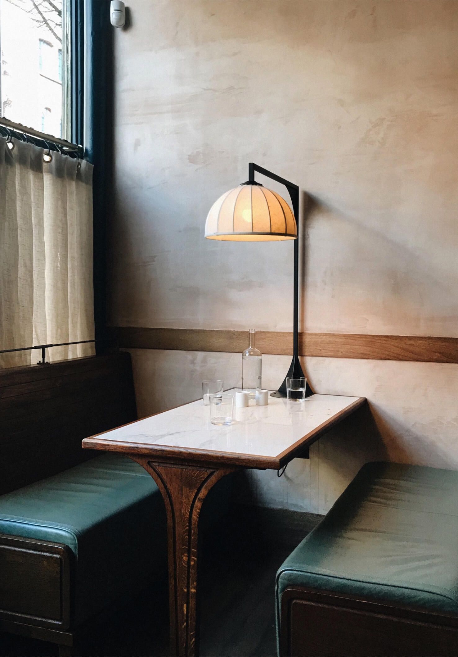 A café corner table with a marble tabletop, dome pendant lamp, dark green banquette seating, and sheer curtains by the window.