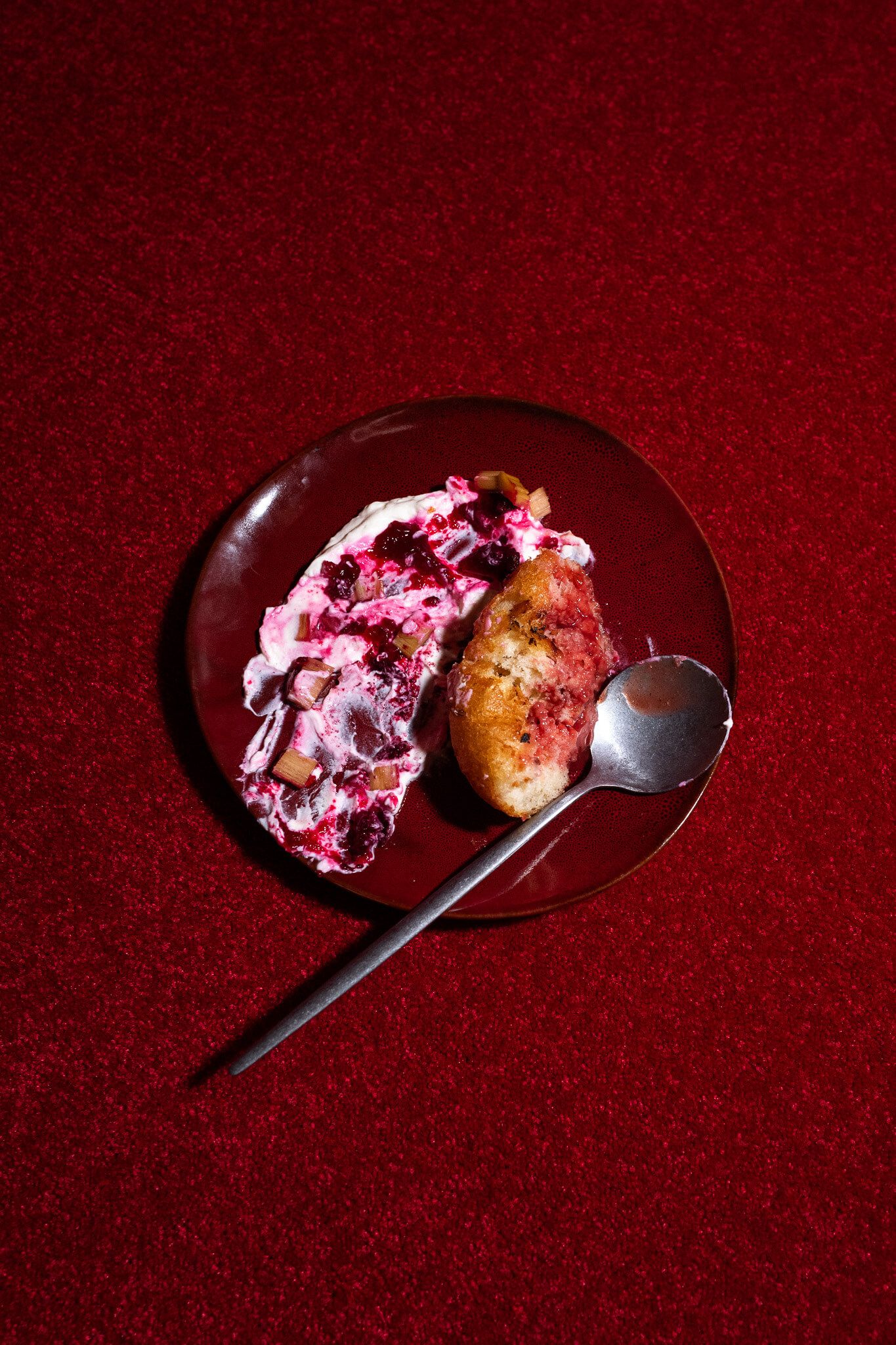 A dessert plate with a berry compote and pastry piece, served with a spoon, on a dark red carpeted surface.