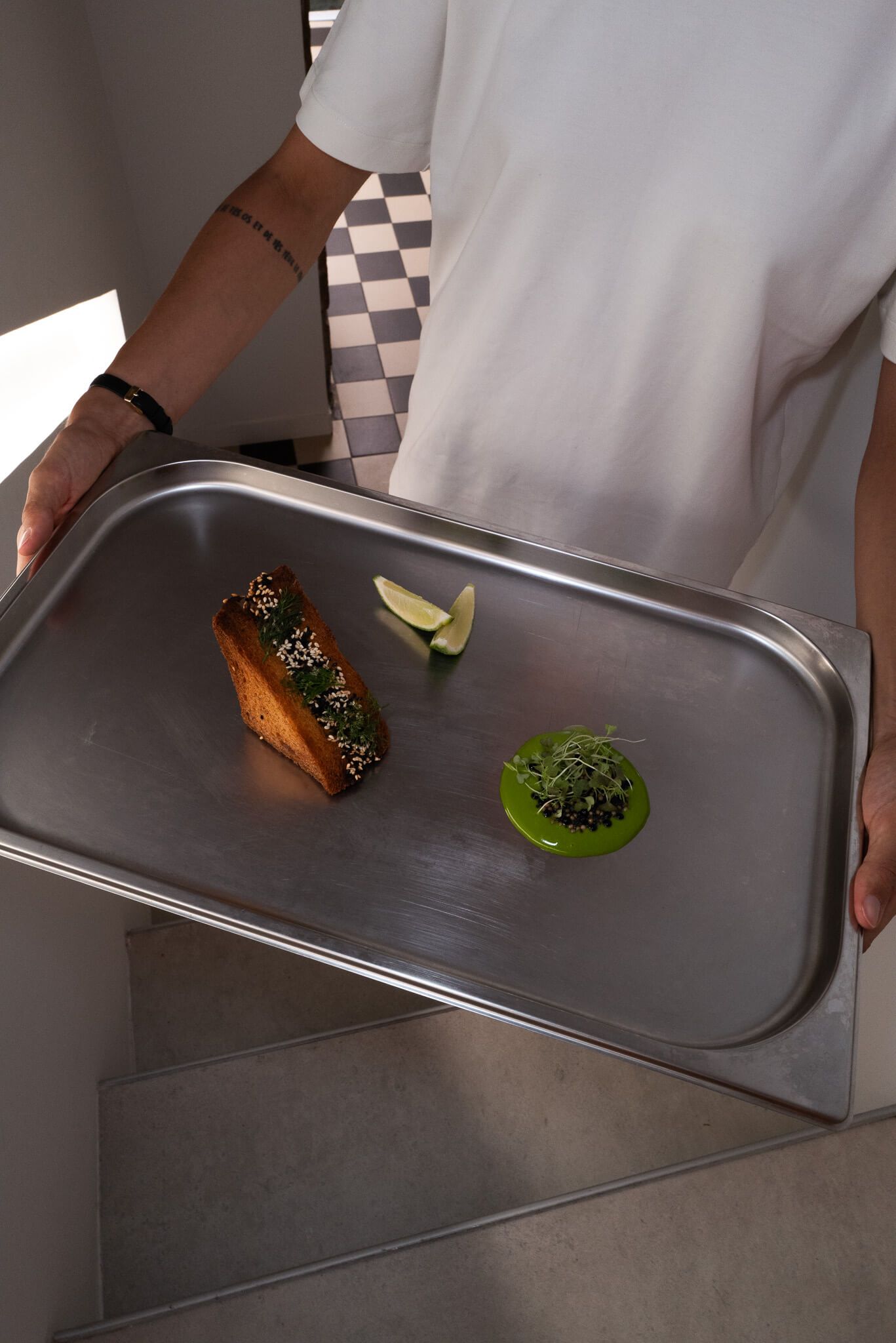 A server presenting a metal tray with a small plated dish, citrus wedge, and green sauce dollop, with a checkered-floor backdrop.