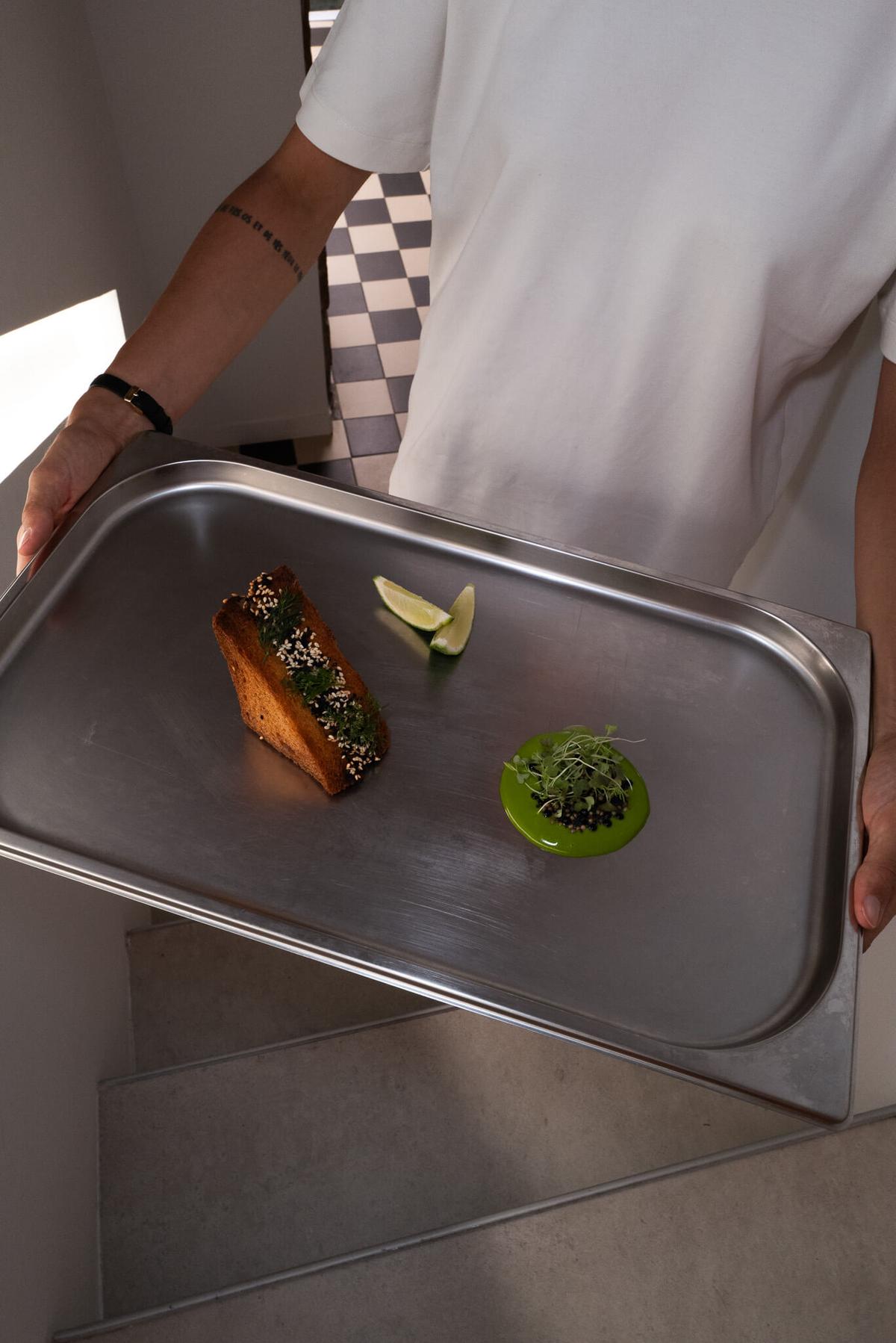 A server presenting a metal tray with a small plated dish, citrus wedge, and green sauce dollop, with a checkered-floor backdrop.