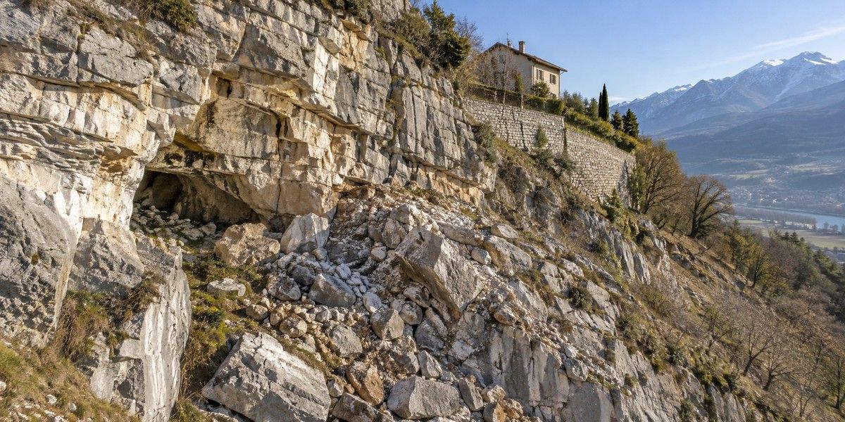 Versant calcaire fracturé avec petite cavité naturelle, blocs rocheux et maison implantée au-dessus d’un mur de soutènement
