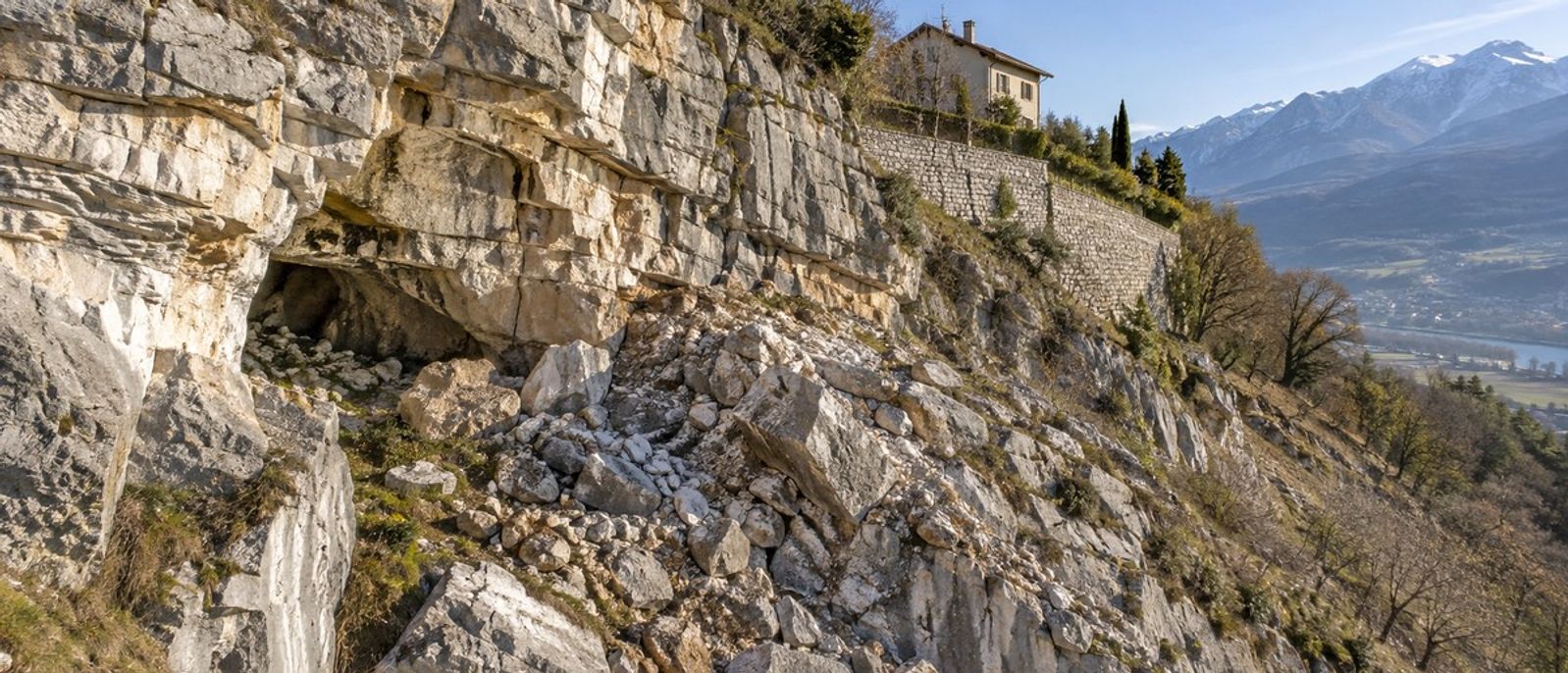 Versant calcaire fracturé avec petite cavité naturelle, blocs rocheux et maison implantée au-dessus d’un mur de soutènement