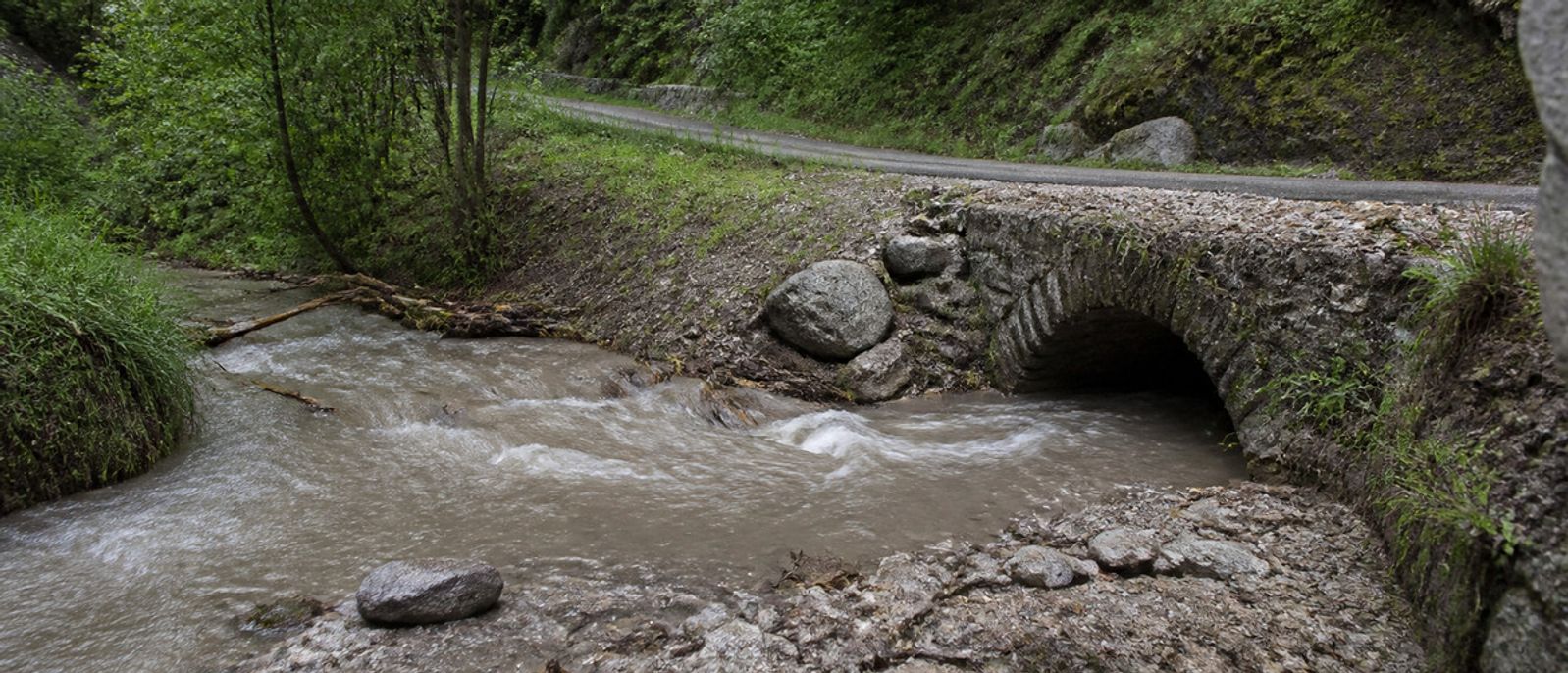 Vallon chargé d’eau boueuse passant sous une petite route de montagne