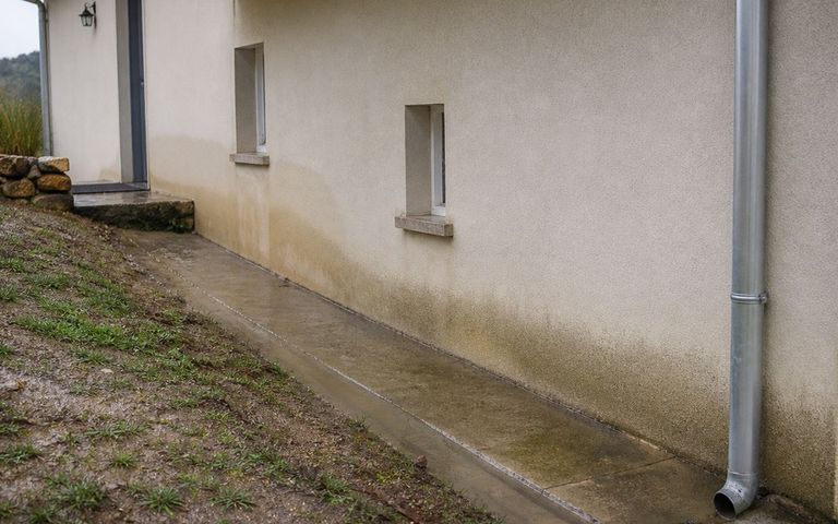 Façade humide d'une maison sur terrain en pente après la pluie, avec descente d'eau et rigole périphérique.