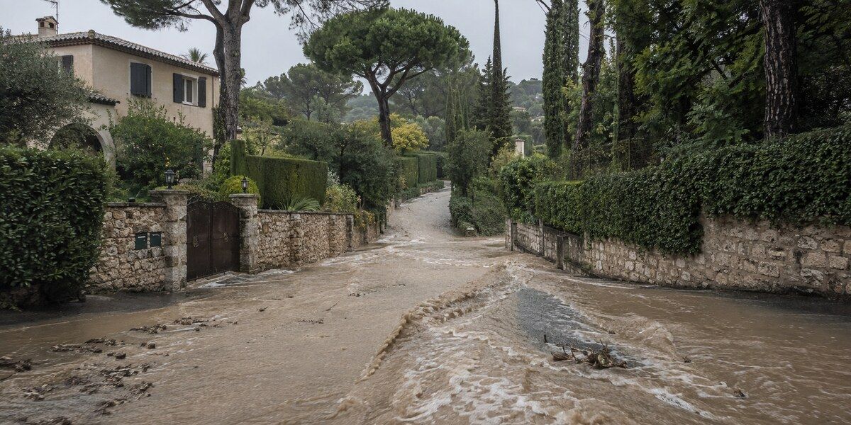Eaux boueuses ruisselant dans une rue résidentielle en pente, bordée de villas et de murs en pierre dans un environnement évoquant Mougins