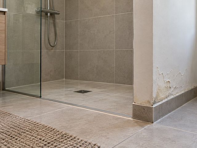 Douche carrelée de plain-pied avec une peinture écaillée et des traces d’humidité au pied de la cloison voisine.