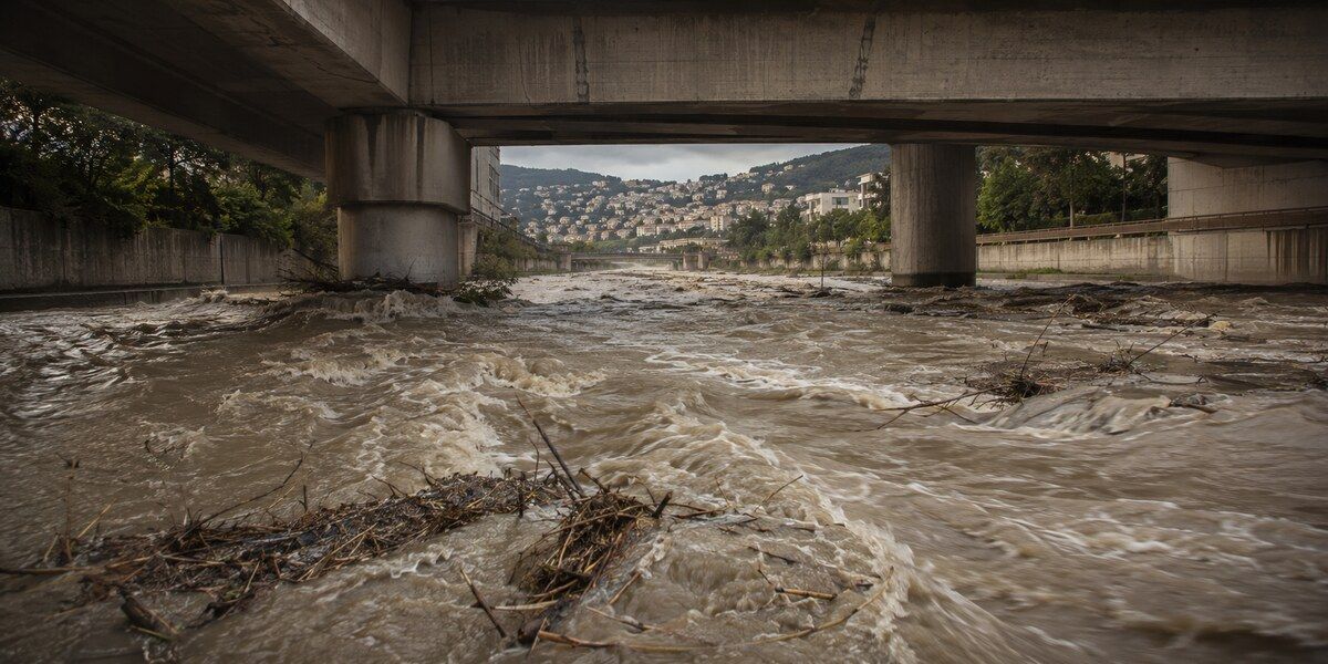 Torrent boueux chargé de branches s’écoulant sous un pont dans un canal urbain évoquant le Paillon à Nice