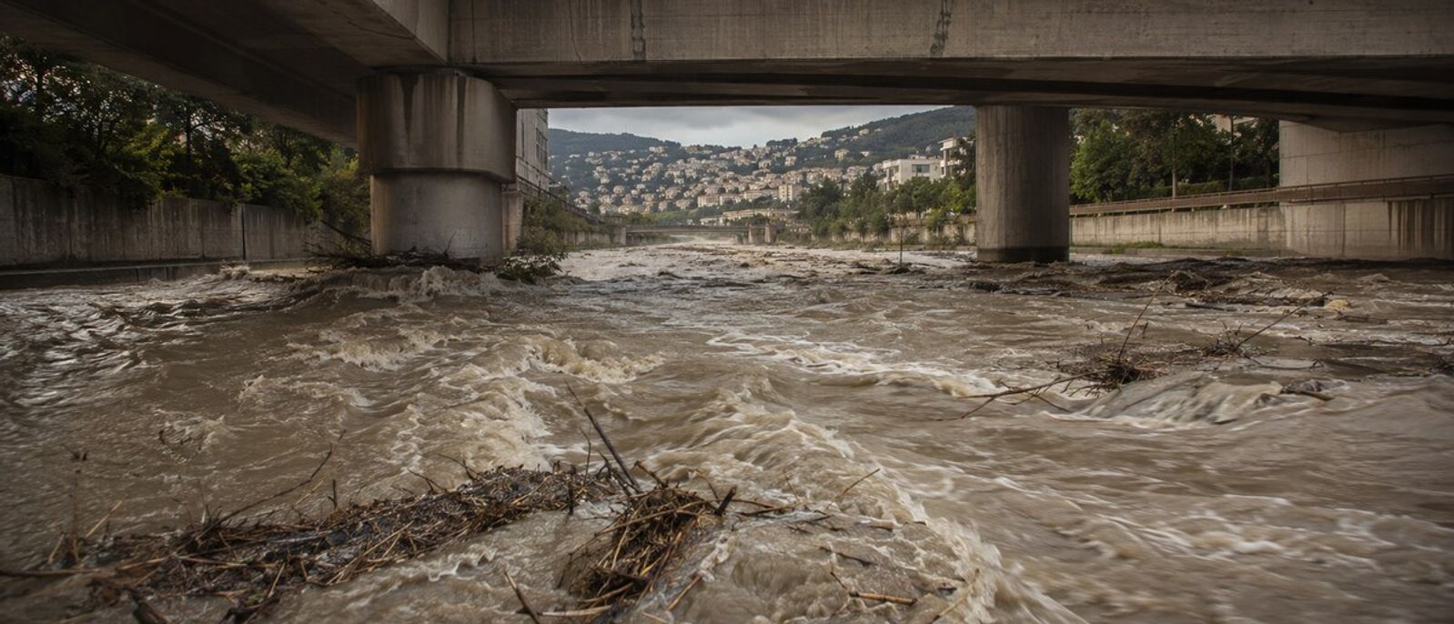 Torrent boueux chargé de branches s’écoulant sous un pont dans un canal urbain évoquant le Paillon à Nice