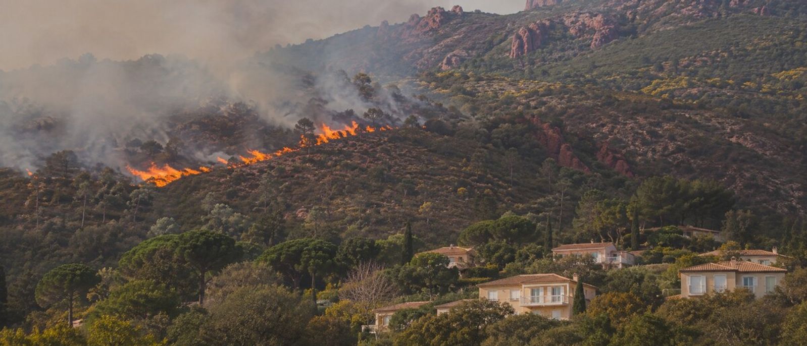 Feu de forêt en journée sur une colline de roches rouges proche de villas et de végétation méditerranéenne évoquant Mandelieu-la-Napoule